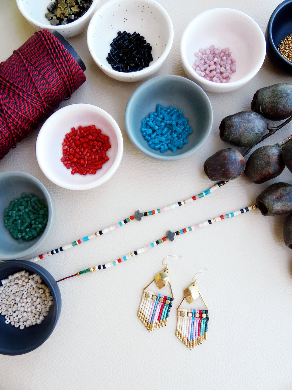 Peut inclure: Un ensemble de perles colorées dans de petits bols, une bobine de fil rouge et deux paires de boucles d'oreilles dorées avec des franges de perles colorées. Les boucles d'oreilles pendent d'une surface blanche.