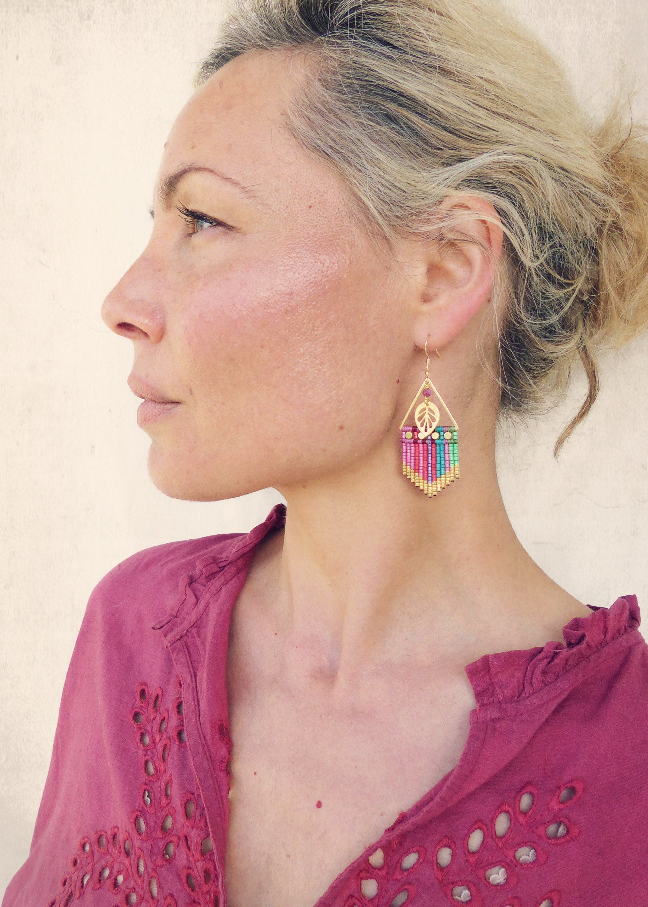 Peut inclure: Une femme portant une chemise rose à bordure en dentelle avec des boucles d'oreilles en perles dorées et multicolores. Les boucles d'oreilles présentent un petit charme en forme de feuille.