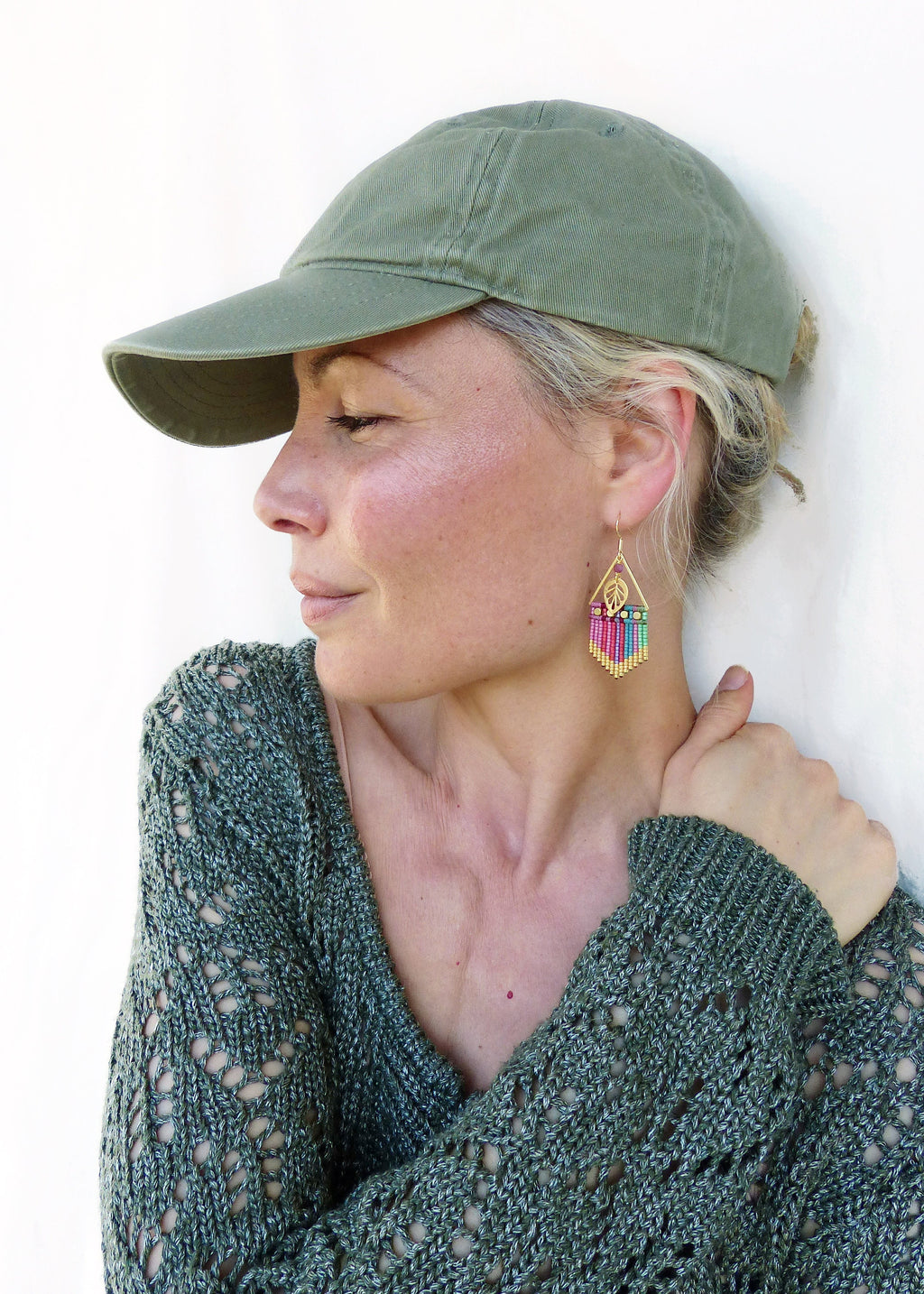 Peut inclure: Une femme portant un pull en tricot vert et une casquette de baseball verte. Elle porte des boucles d'oreilles en perles colorées avec un motif de feuille d'or.