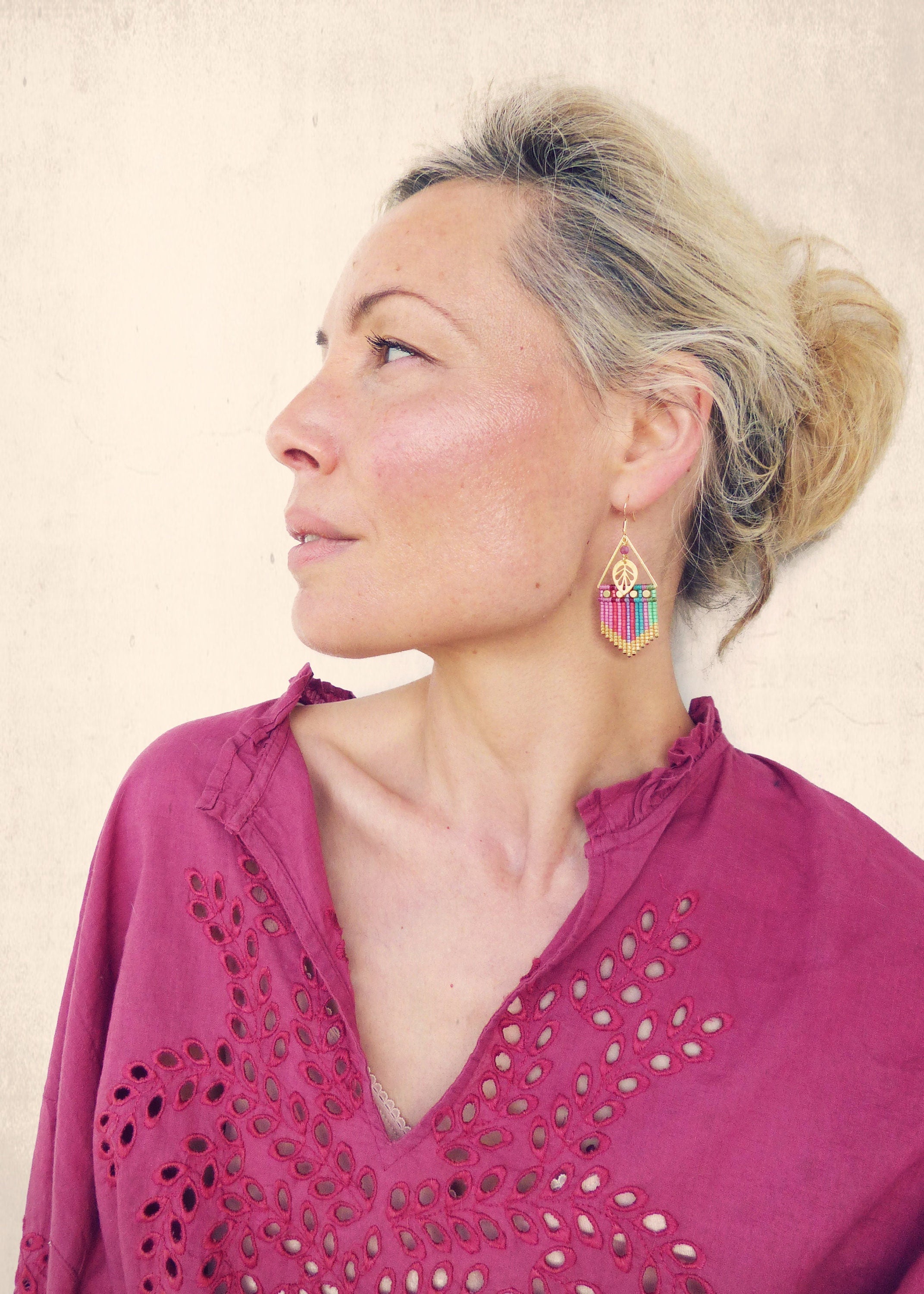 Peut inclure: Une femme portant un haut rose brodé avec une paire de boucles d'oreilles en perles colorées. Les boucles d'oreilles sont dorées et présentent un charme en forme de signe de paix.