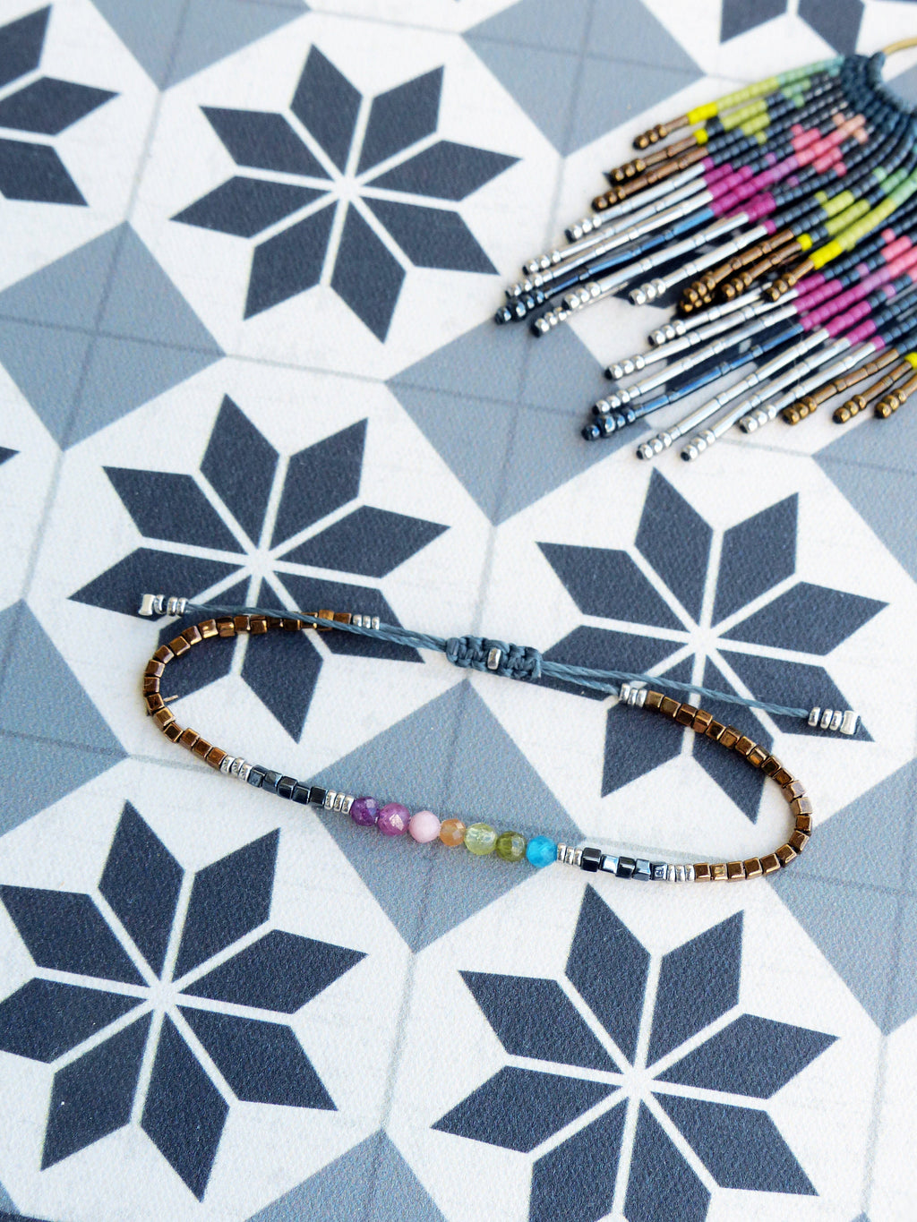 Peut inclure: Un bracelet en perles avec une variété de pierres colorées et des perles argentées et cuivrées. Le bracelet est posé sur une surface carrelée blanche et grise.