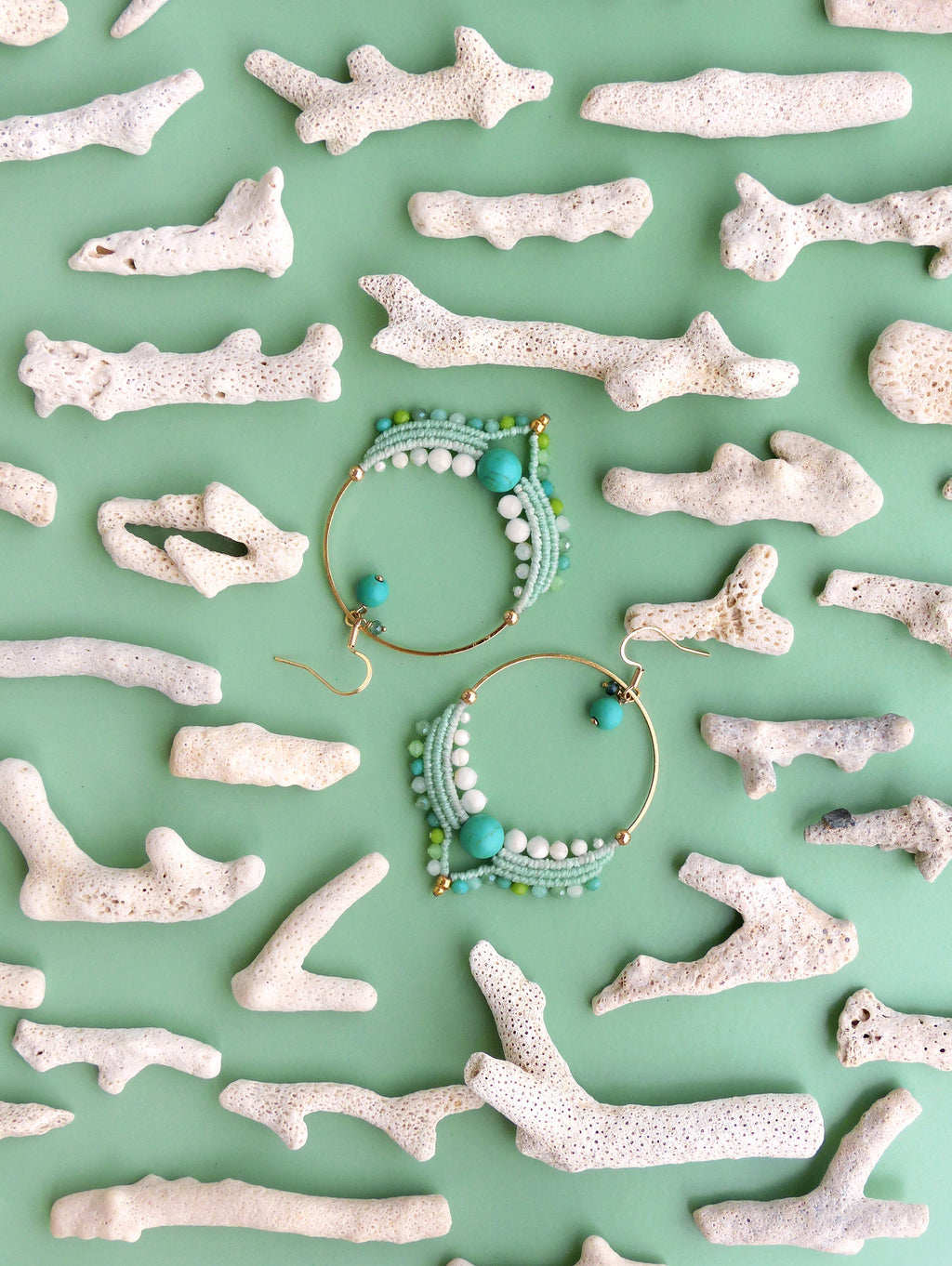 Peut inclure: Deux boucles d'oreilles créoles dorées avec des perles turquoises, blanches et vertes, et des détails en macramé. Les boucles d'oreilles sont présentées sur une surface vert clair, entourées de morceaux de corail blanc.