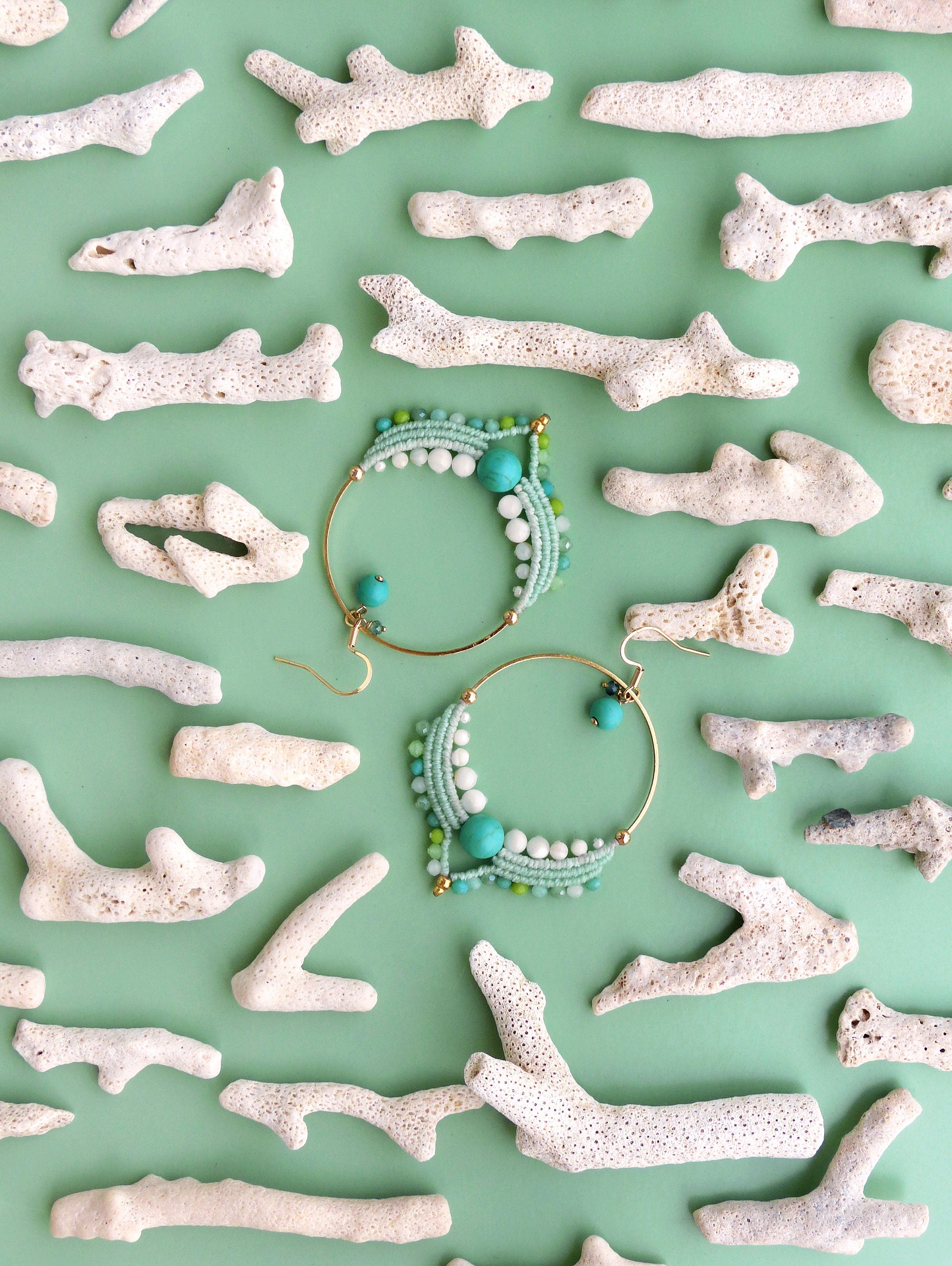 Peut inclure: Deux boucles d'oreilles créoles dorées avec des perles turquoises, blanches et vertes, et des détails en macramé. Les boucles d'oreilles sont présentées sur une surface vert clair, entourées de morceaux de corail blanc.