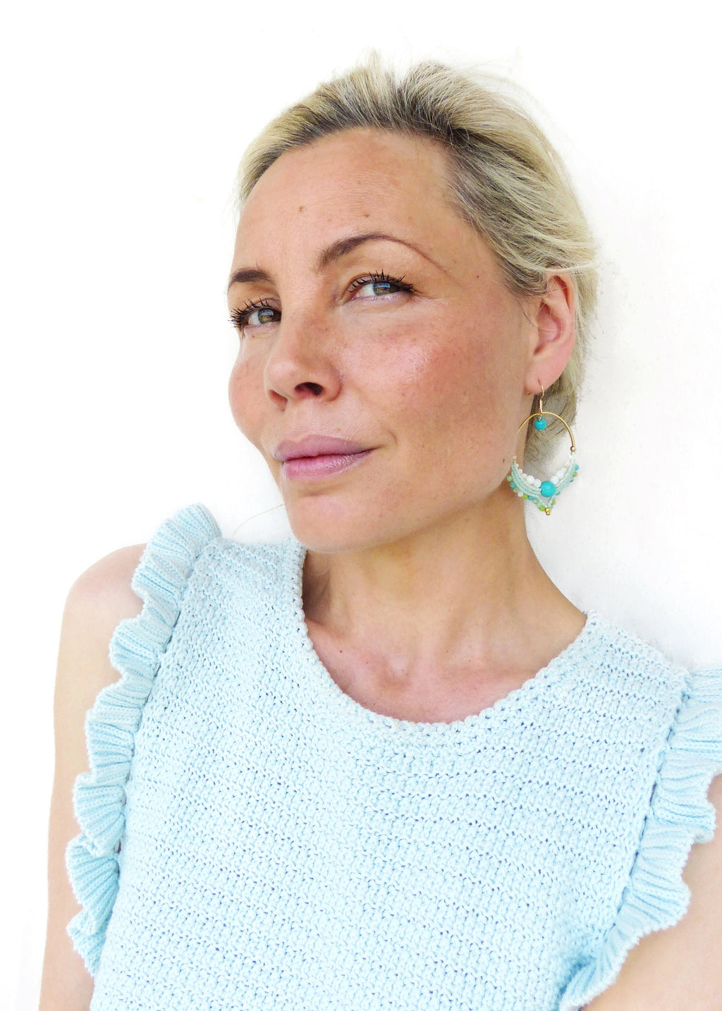 Peut inclure: Une femme porte des boucles d'oreilles dorées avec des perles turquoise et blanches. Les boucles d'oreilles sont au centre de l'attention, contrastant avec son haut en tricot bleu clair à manches volantées. Le fond est blanc uni.