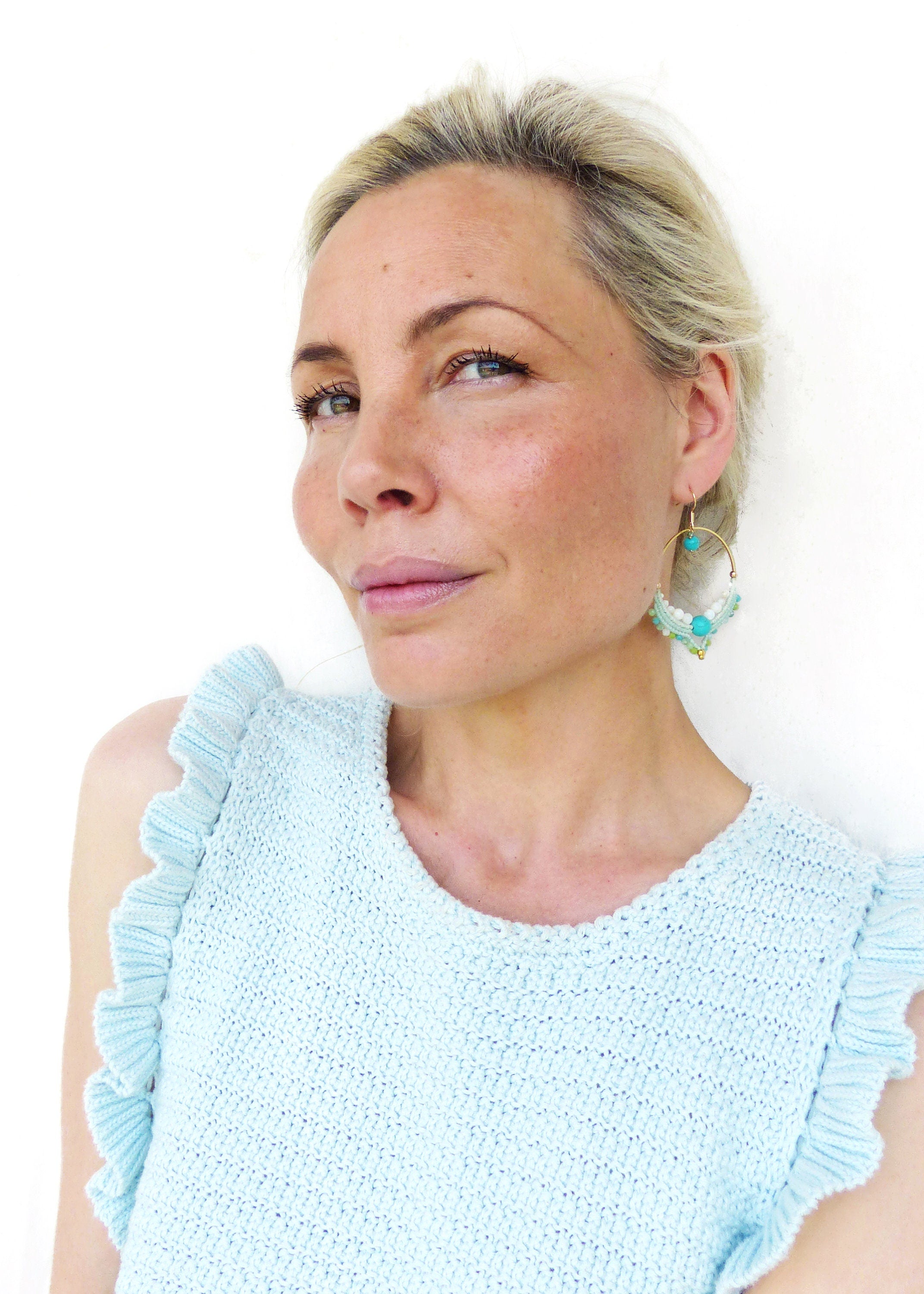 Peut inclure: Une femme porte des boucles d'oreilles dorées avec des perles turquoise et blanches. Les boucles d'oreilles sont au centre de l'attention, contrastant avec son haut en tricot bleu clair à manches volantées. Le fond est blanc uni.