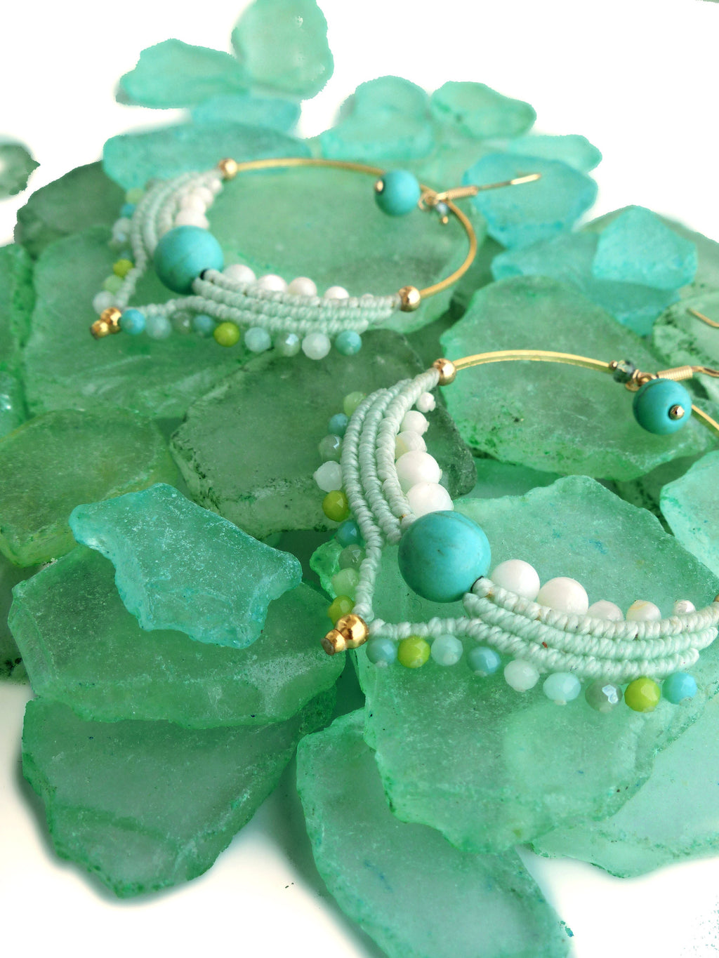 Peut inclure: Une paire de boucles d'oreilles créoles faites à la main avec un design bohème. Les boucles d'oreilles présentent un cerceau en métal doré, orné de macramé bleu clair, de perles blanches, de perles turquoise et de petites perles vertes. Les boucles d'oreilles sont présentées sur un lit de verre de mer vert.