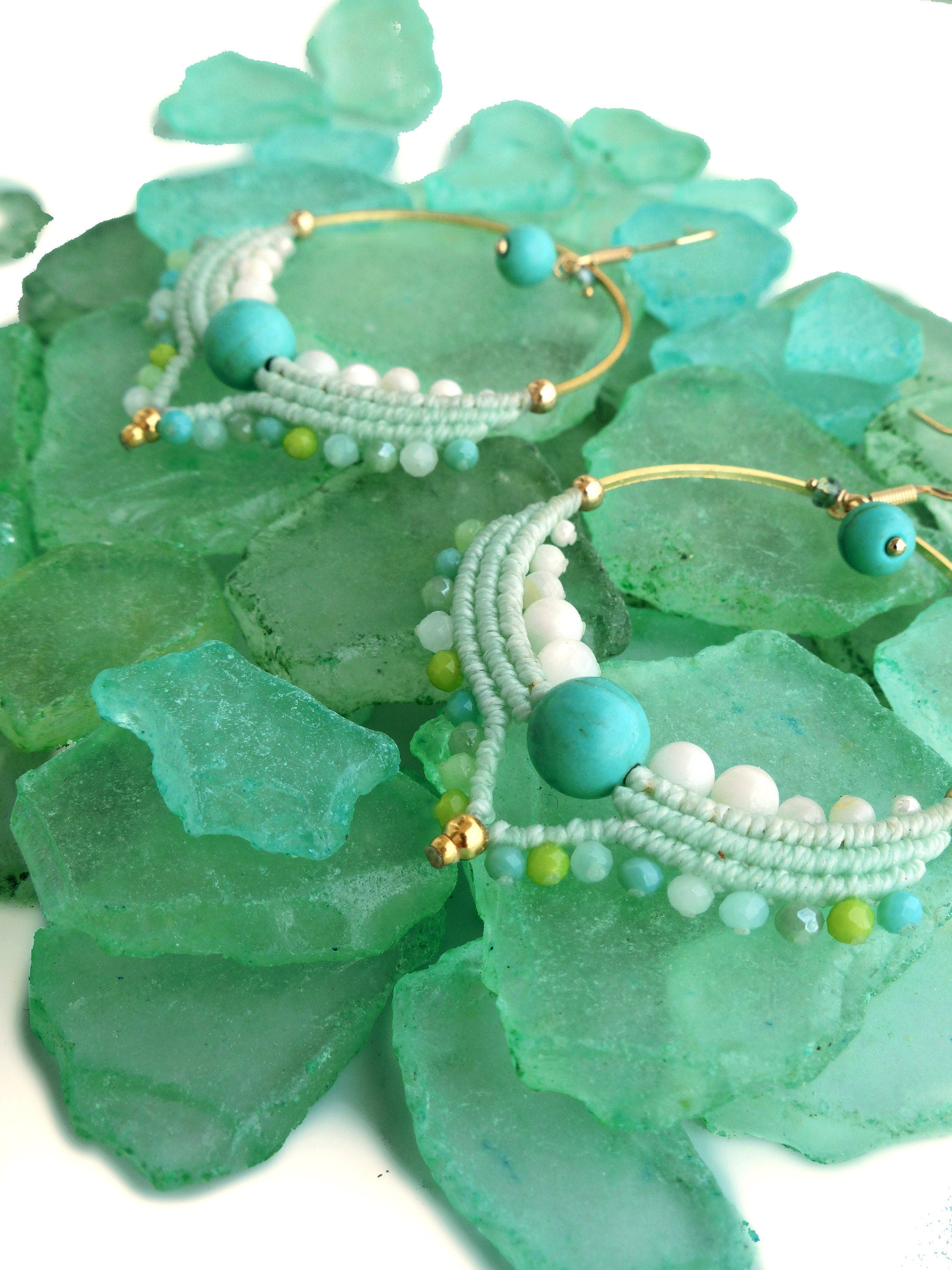 Peut inclure: Une paire de boucles d'oreilles créoles faites à la main avec un design bohème. Les boucles d'oreilles présentent un cerceau en métal doré, orné de macramé bleu clair, de perles blanches, de perles turquoise et de petites perles vertes. Les boucles d'oreilles sont présentées sur un lit de verre de mer vert.