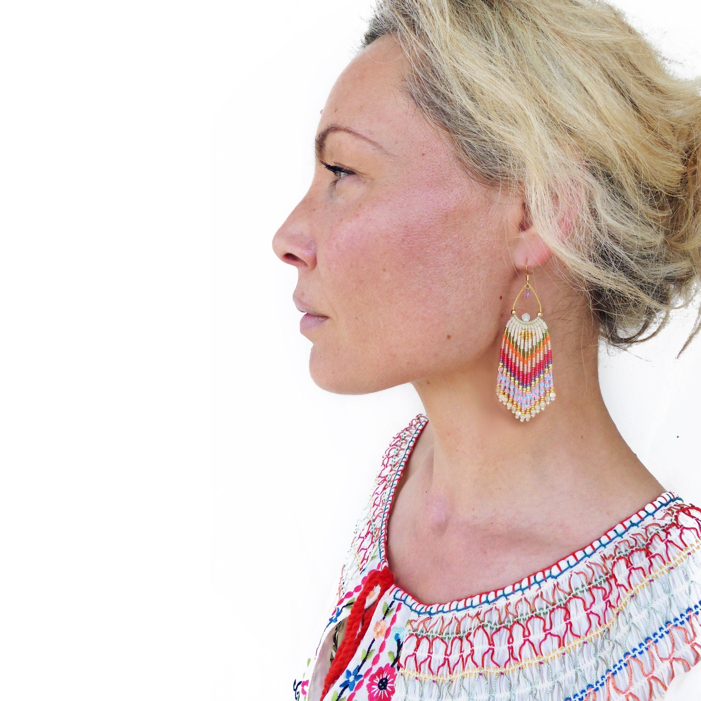 Peut inclure: Une femme portant un haut blanc brodé de motifs floraux colorés et de longues boucles d'oreilles en perles avec un motif géométrique dans des tons de rouge, orange, rose, violet et blanc.