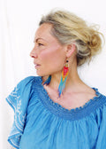 Peut inclure: Une femme portant un haut en jean bleu décolleté avec des broderies blanches et de longues boucles d'oreilles en perles colorées.