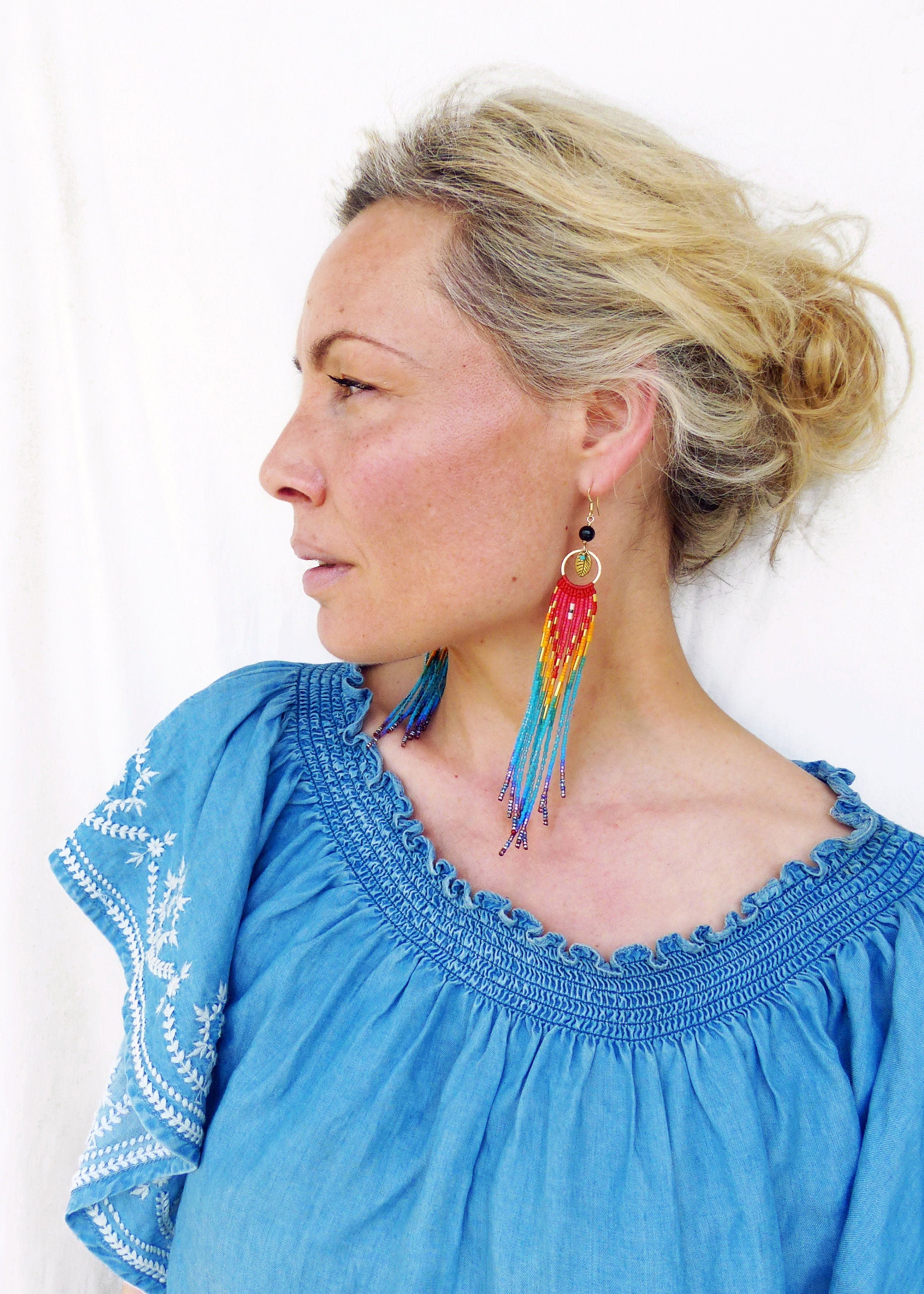 Peut inclure: Une femme portant un haut en jean bleu décolleté avec des broderies blanches et de longues boucles d'oreilles en perles colorées.