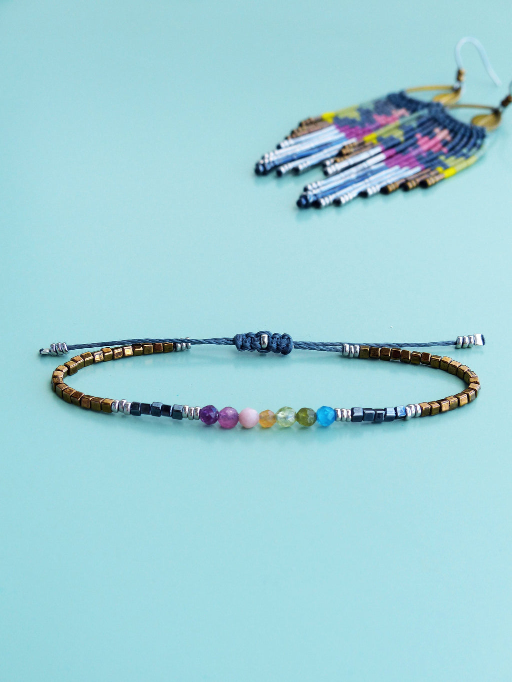 Peut inclure: Un bracelet délicat avec des perles de pierres précieuses arc-en-ciel et des accents dorés et argentés. Le bracelet est fait avec un cordon gris et a une fermeture à nœud coulissant.