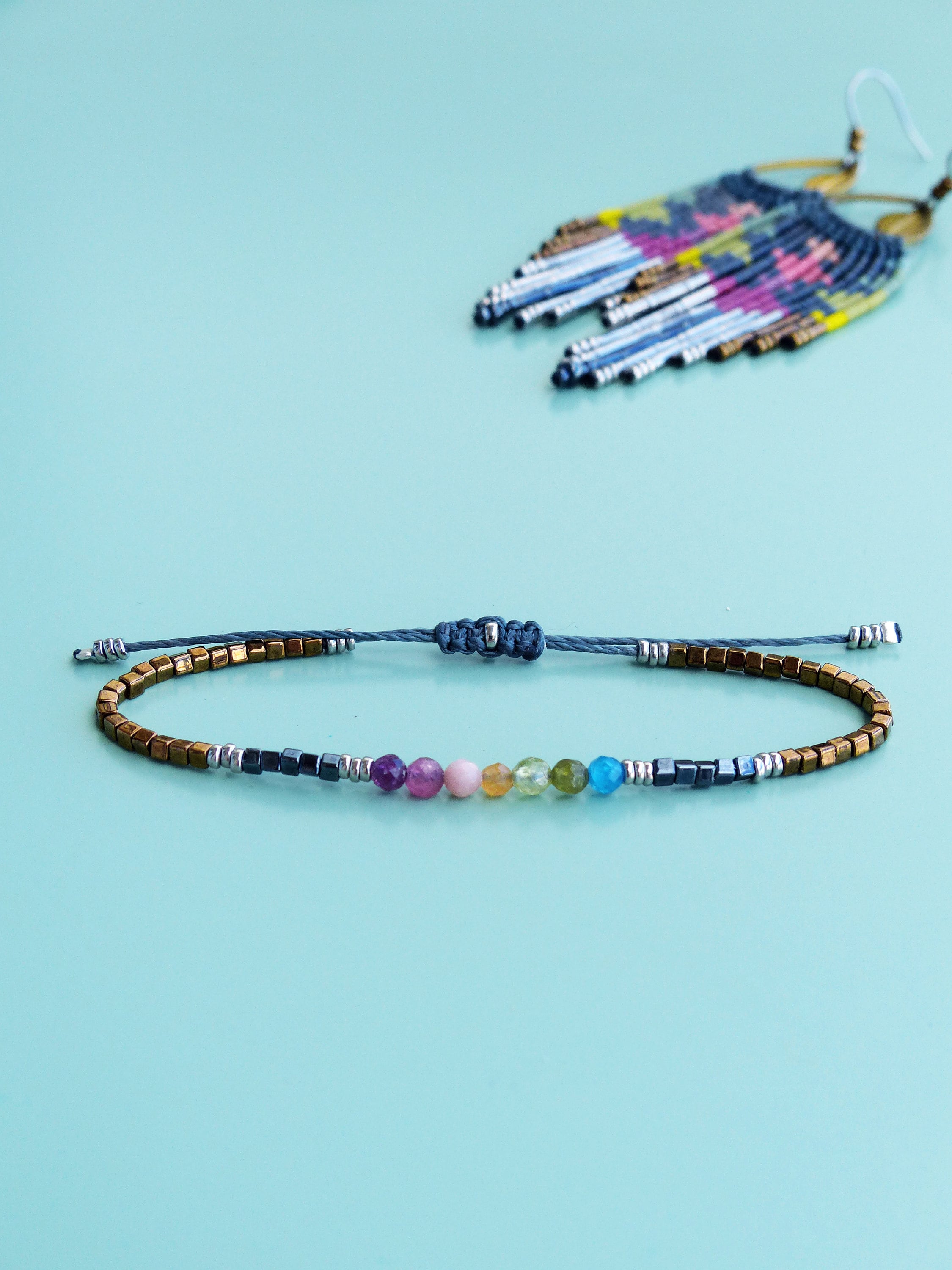 Peut inclure: Un bracelet délicat avec des perles de pierres précieuses arc-en-ciel et des accents dorés et argentés. Le bracelet est fait avec un cordon gris et a une fermeture à nœud coulissant.