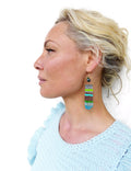 Peut inclure: Une femme portant un pull bleu clair avec une paire de longues boucles d'oreilles en perles colorées. Les boucles d'oreilles sont faites de petites perles colorées et ont un anneau en métal doré en haut.