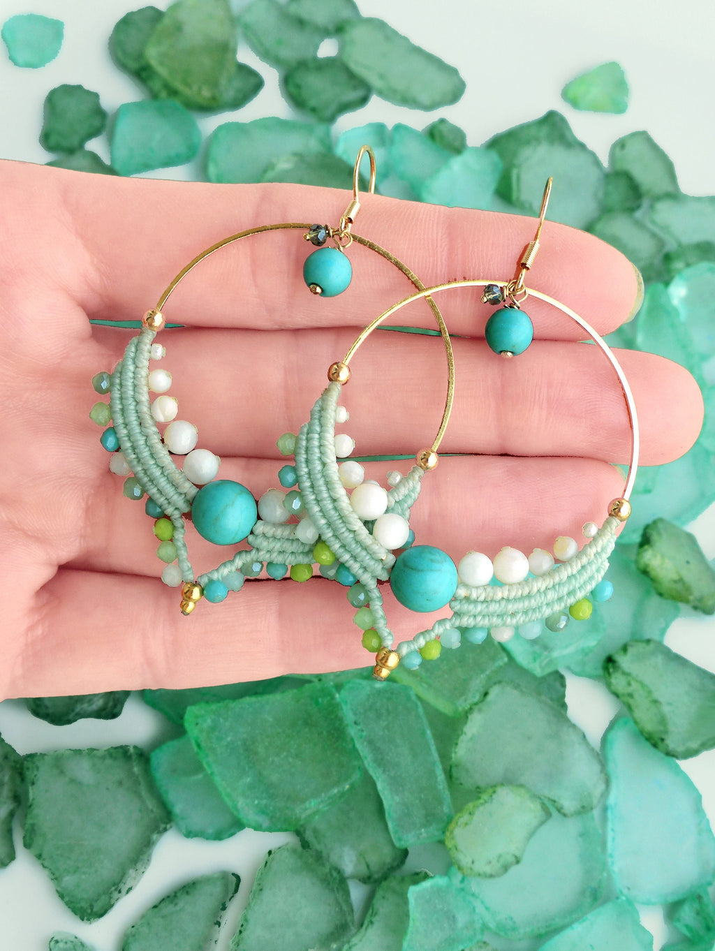 Peut inclure: Boucles d'oreilles créoles dorées ornées de perles turquoise, blanches et vertes. Les boucles d'oreilles présentent un motif macramé avec une perle turquoise au centre et de petites perles de différentes nuances de vert et de blanc. Fond de verre de mer vert.