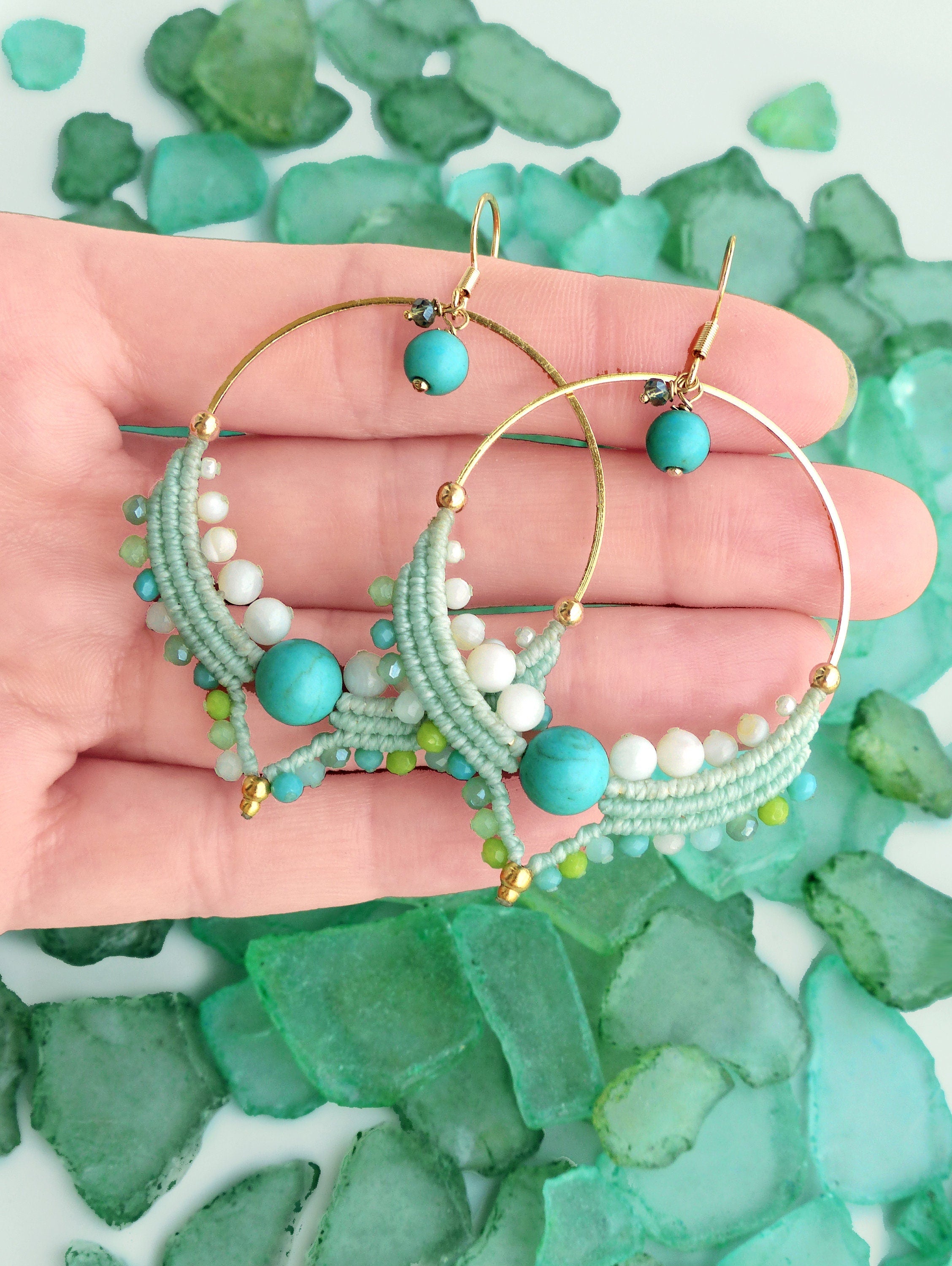 Peut inclure: Boucles d'oreilles créoles dorées ornées de perles turquoise, blanches et vertes. Les boucles d'oreilles présentent un motif macramé avec une perle turquoise au centre et de petites perles de différentes nuances de vert et de blanc. Fond de verre de mer vert.