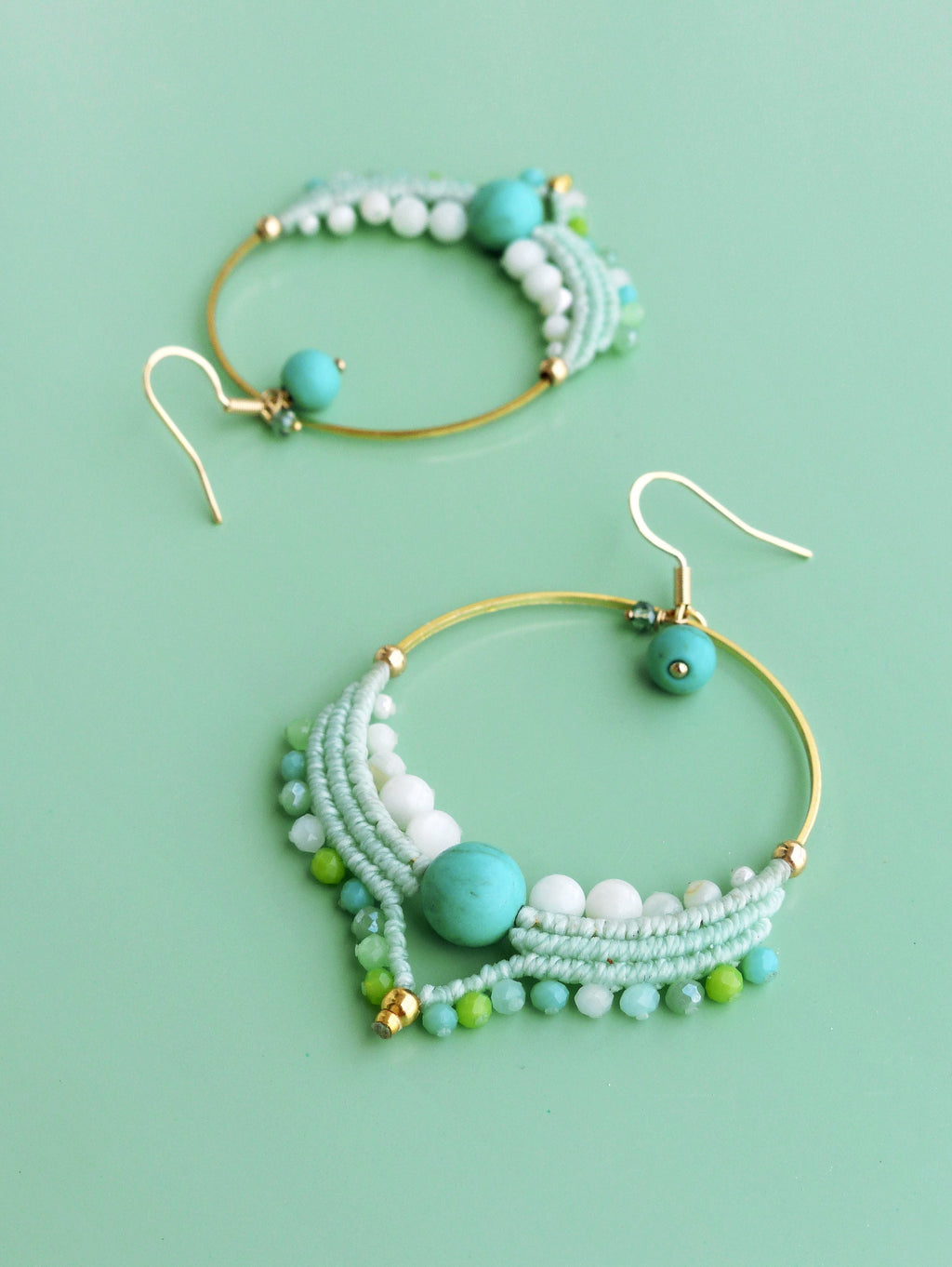Peut inclure: Deux boucles d'oreilles créoles dorées ornées de perles turquoise, blanches et vertes. Les boucles d'oreilles présentent un motif macramé avec une perle turquoise au centre et un crochet doré. Fond vert clair.