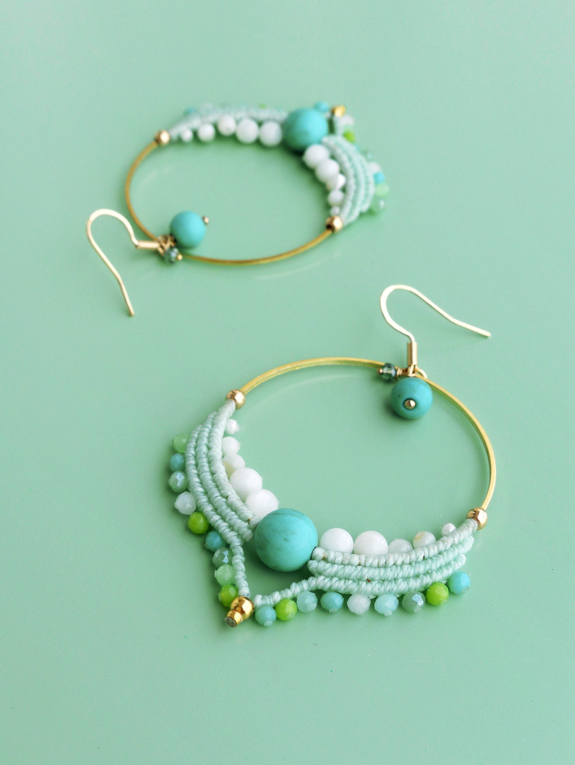 Peut inclure: Deux boucles d'oreilles créoles dorées ornées de perles turquoise, blanches et vertes. Les boucles d'oreilles présentent un motif macramé avec une perle turquoise au centre et un crochet doré. Fond vert clair.