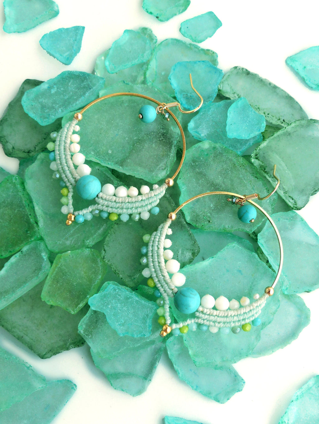 Peut inclure: Boucles d'oreilles créoles dorées avec des perles turquoises et blanches, et des détails en macramé. Les boucles d'oreilles sont posées sur un fond de verre de mer vert et bleu. Les boucles d'oreilles ont un style bohème.