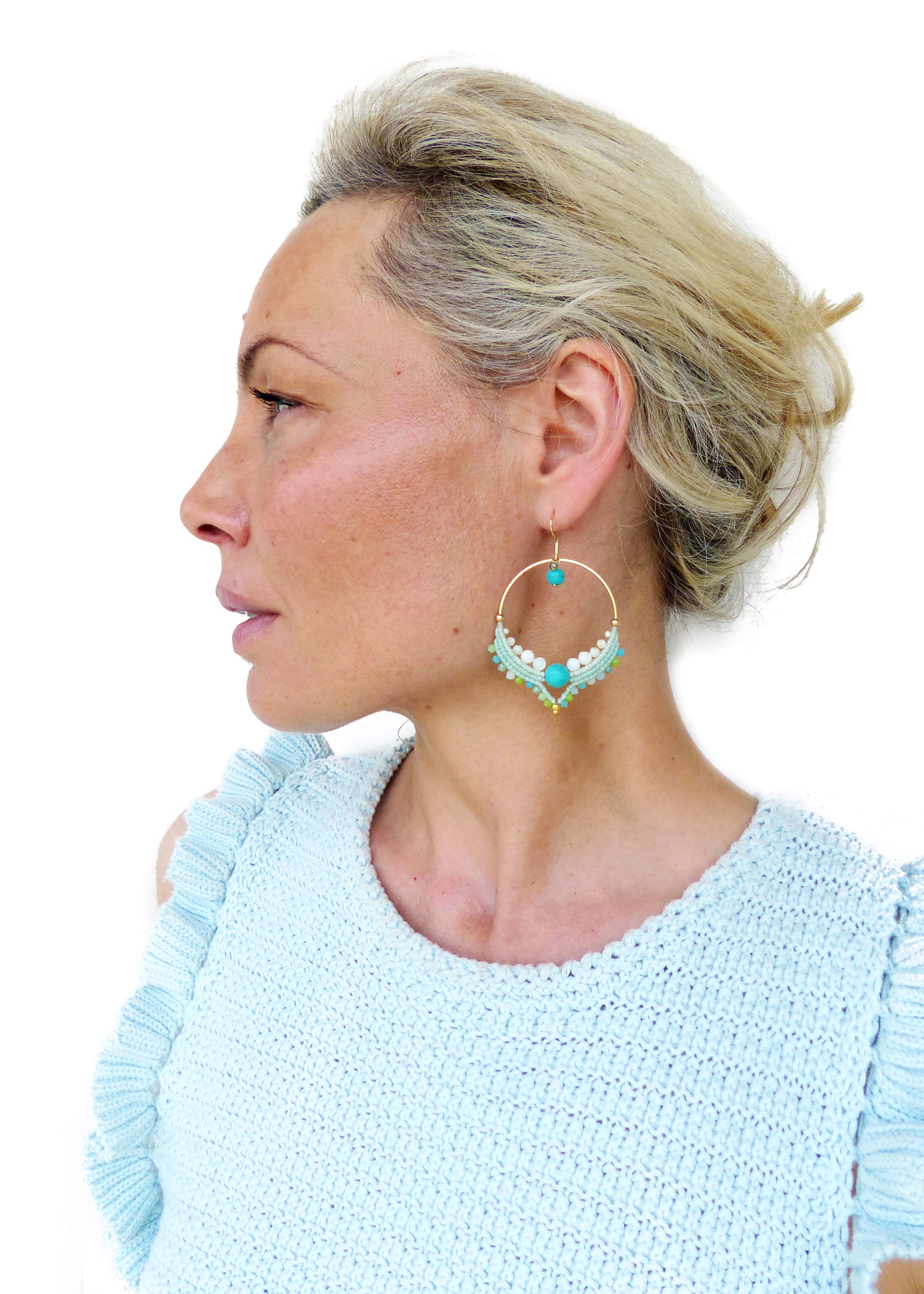 Peut inclure: Boucles d'oreilles créoles dorées avec des perles turquoise et blanches. Les boucles d'oreilles présentent un design macramé avec une pierre turquoise au centre et une petite pierre turquoise au sommet de la créole. La personne porte un haut bleu clair.