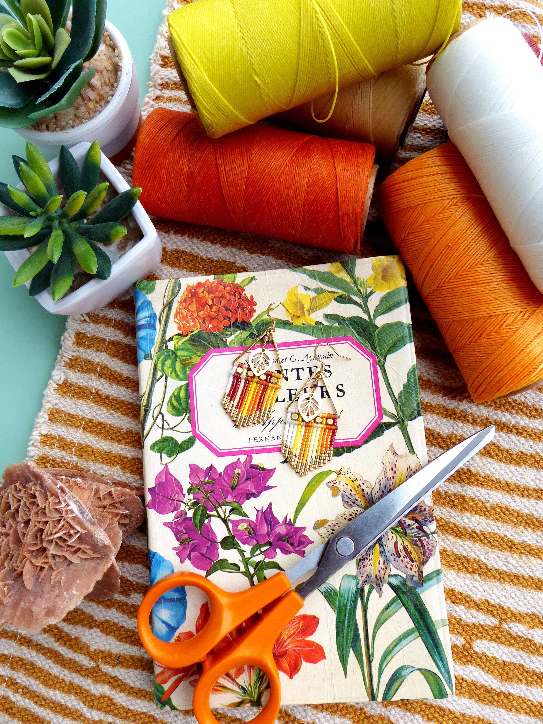 Peut inclure: Une paire de boucles d'oreilles en perles colorées avec un motif floral, une paire de ciseaux orange et des bobines de fil orange, jaune et blanc sur un fond de tissu rayé. Les boucles d'oreilles sont sur un livre avec une couverture florale et le texte "Winter Leaves" sur le dos.