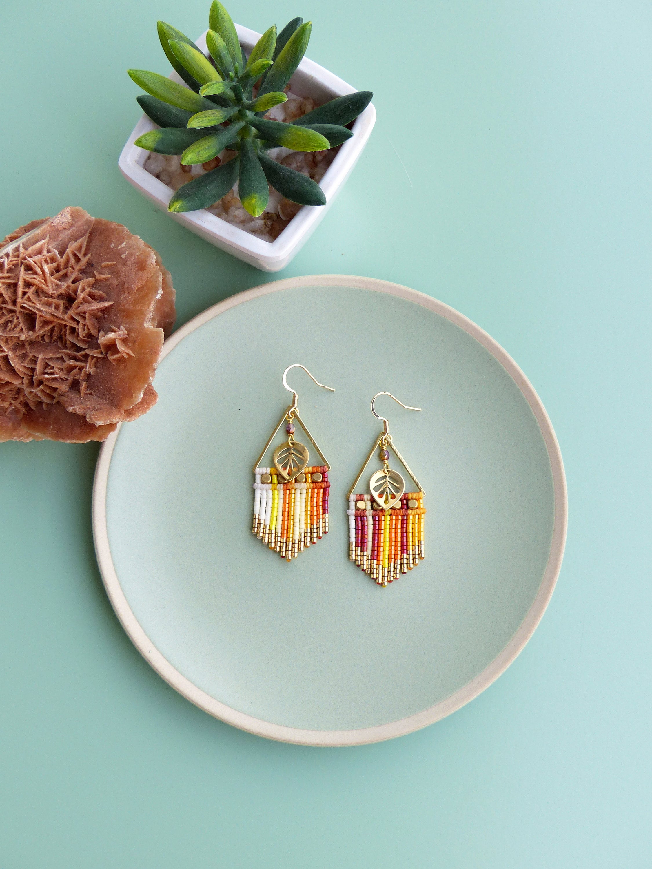 Peut inclure: Une paire de boucles d'oreilles pendantes dorées avec un charme en forme de feuille et une frange de perles colorées. Les boucles d'oreilles sont présentées sur une assiette vert clair.