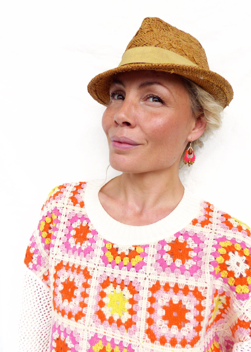 Peut inclure: Une femme portant un pull en crochet blanc avec un motif géométrique coloré. Elle porte un chapeau de paille brun avec un bandeau jaune et des boucles d'oreilles roses.
