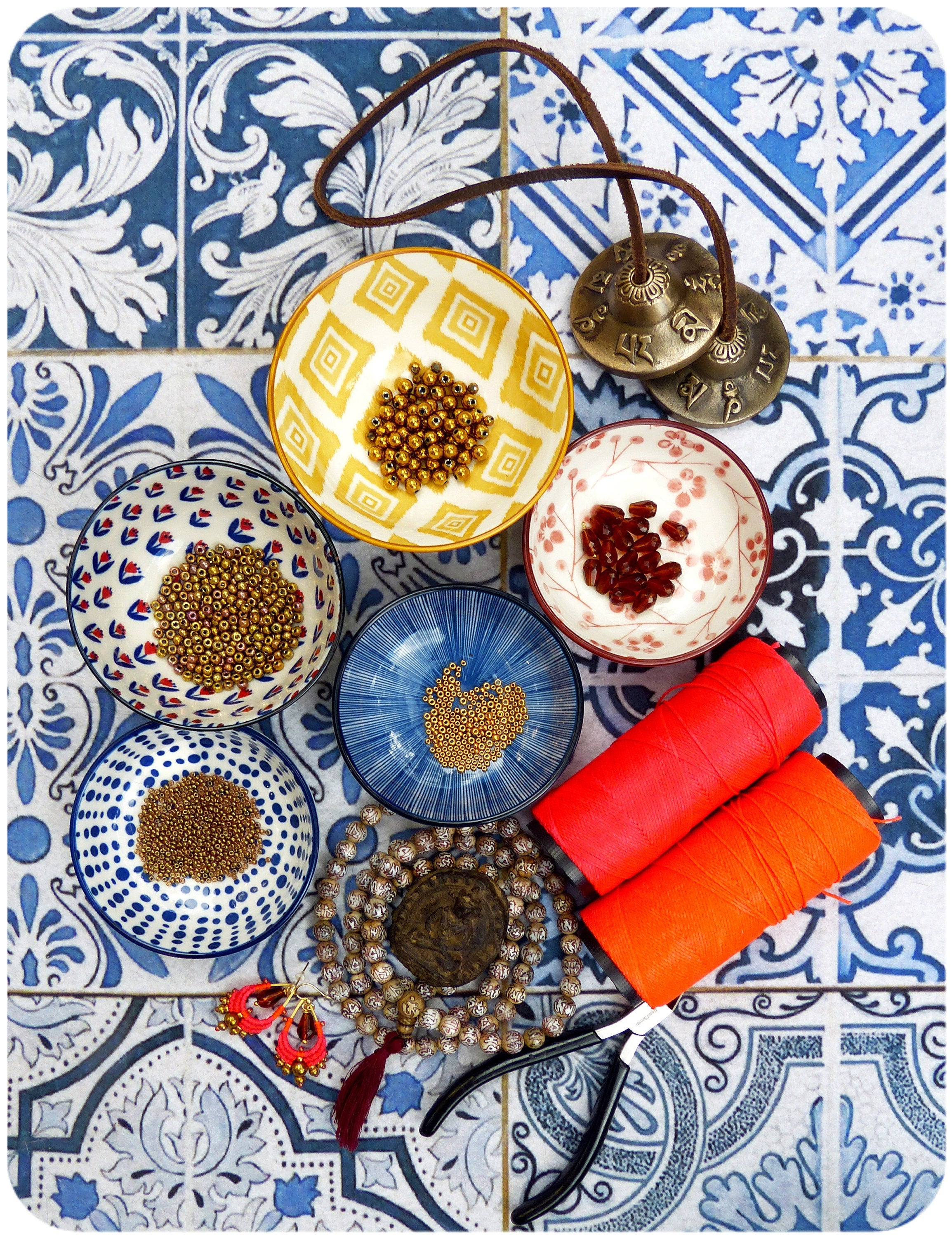 Peut inclure: Assortiment de fournitures artisanales comprenant de petits bols remplis de perles, un collier mala en bois, des bobines de fil rouge et orange, une pince et deux petites cymbales en laiton.