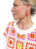 Peut inclure: Une femme portant un pull en crochet blanc avec un motif de carrés de grand-mère colorés. Le pull est de couleur rose, orange et jaune. Elle porte des boucles d'oreilles pendantes roses et dorées.