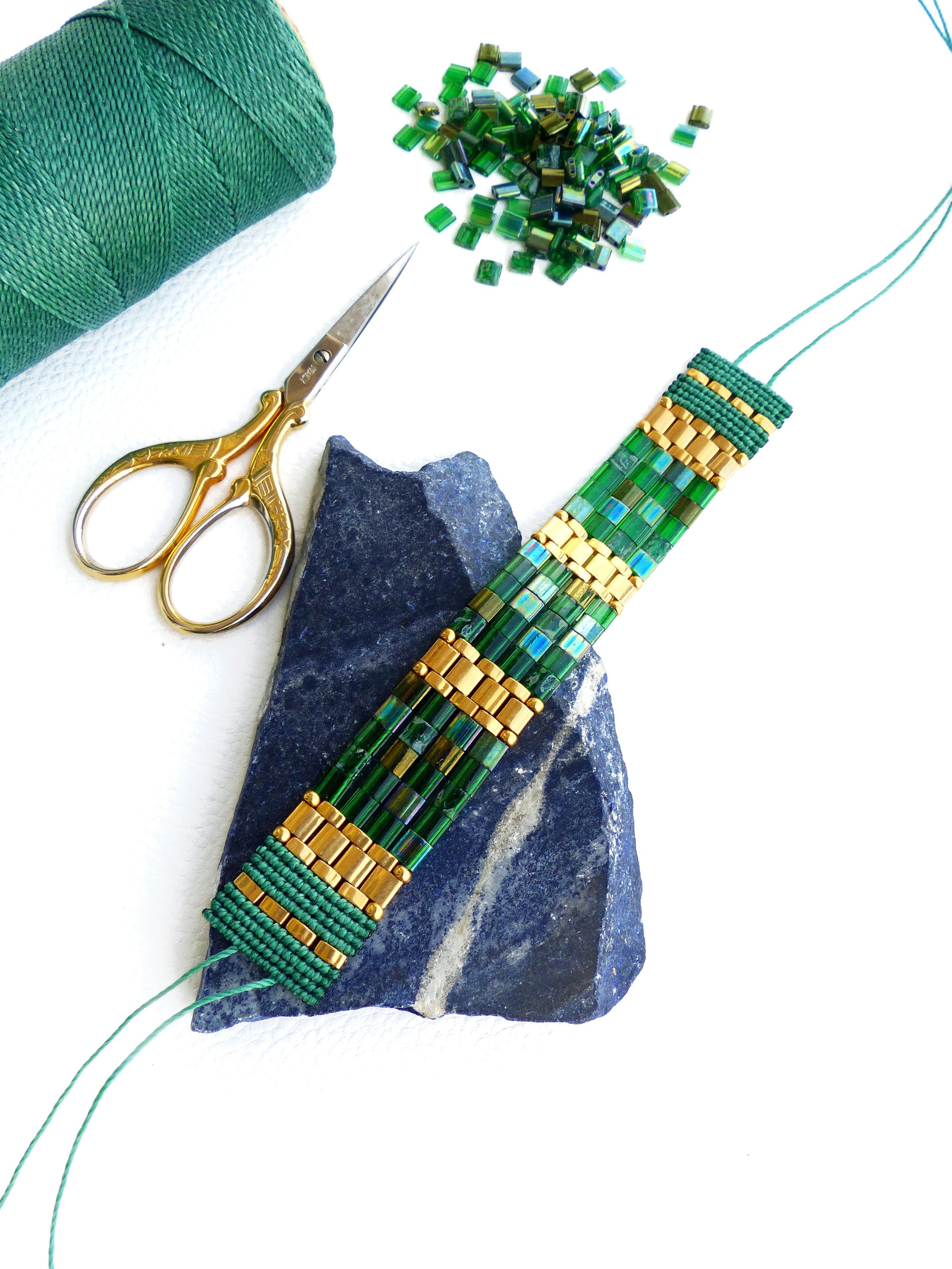 Peut inclure: Un bracelet en perles vertes et dorées sur une pierre bleu-gris avec du fil vert et des ciseaux. Il y a un tas de perles vertes en arrière-plan.