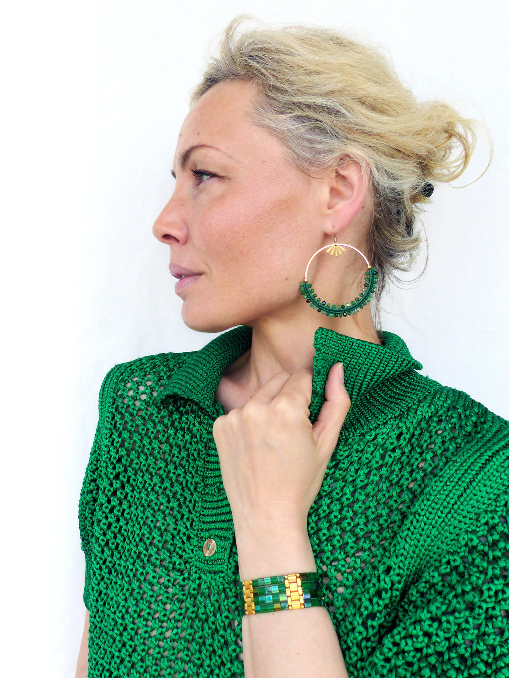 Peut inclure: Une femme portant un pull en tricot vert avec un bracelet en perles vertes et dorées au poignet. Elle porte également des boucles d'oreilles créoles en perles vertes et dorées.