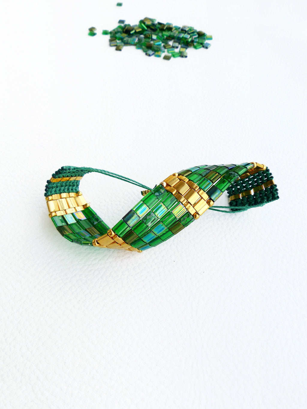 Peut inclure: Un bracelet en perles vertes et dorées avec un design torsadé. Le bracelet est fait de petites perles carrées et est posé sur une surface blanche.