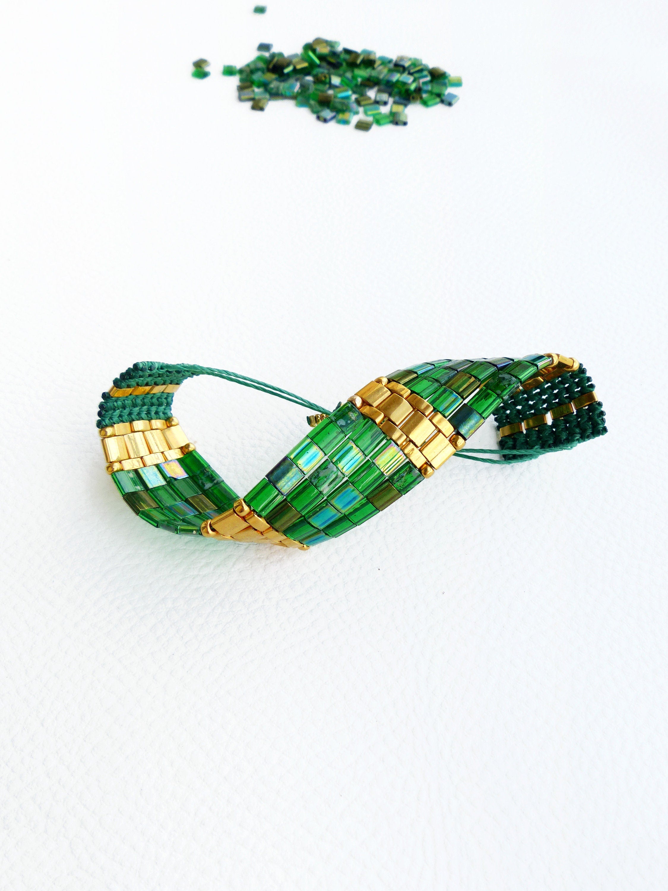 Peut inclure: Un bracelet en perles vertes et dorées avec un design torsadé. Le bracelet est fait de petites perles carrées et est posé sur une surface blanche.