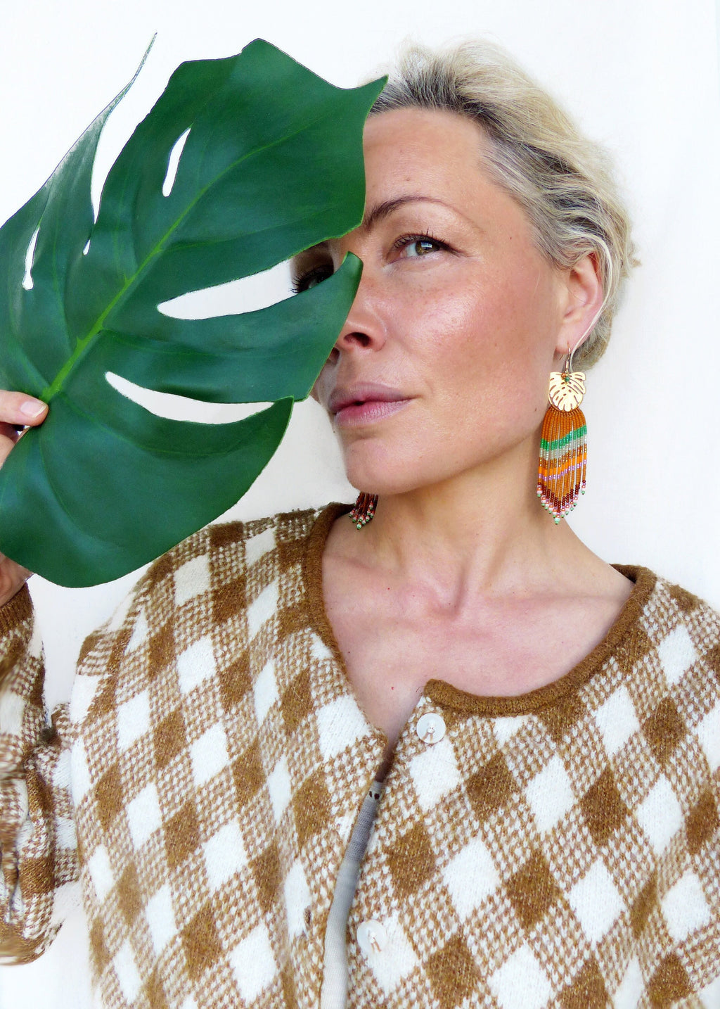 Peut inclure: Boucles d'oreilles dorées en forme de feuille avec des franges de perles colorées. Les boucles d'oreilles sont associées à un cardigan à carreaux marron et blanc. Une grande feuille verte masque partiellement le visage.