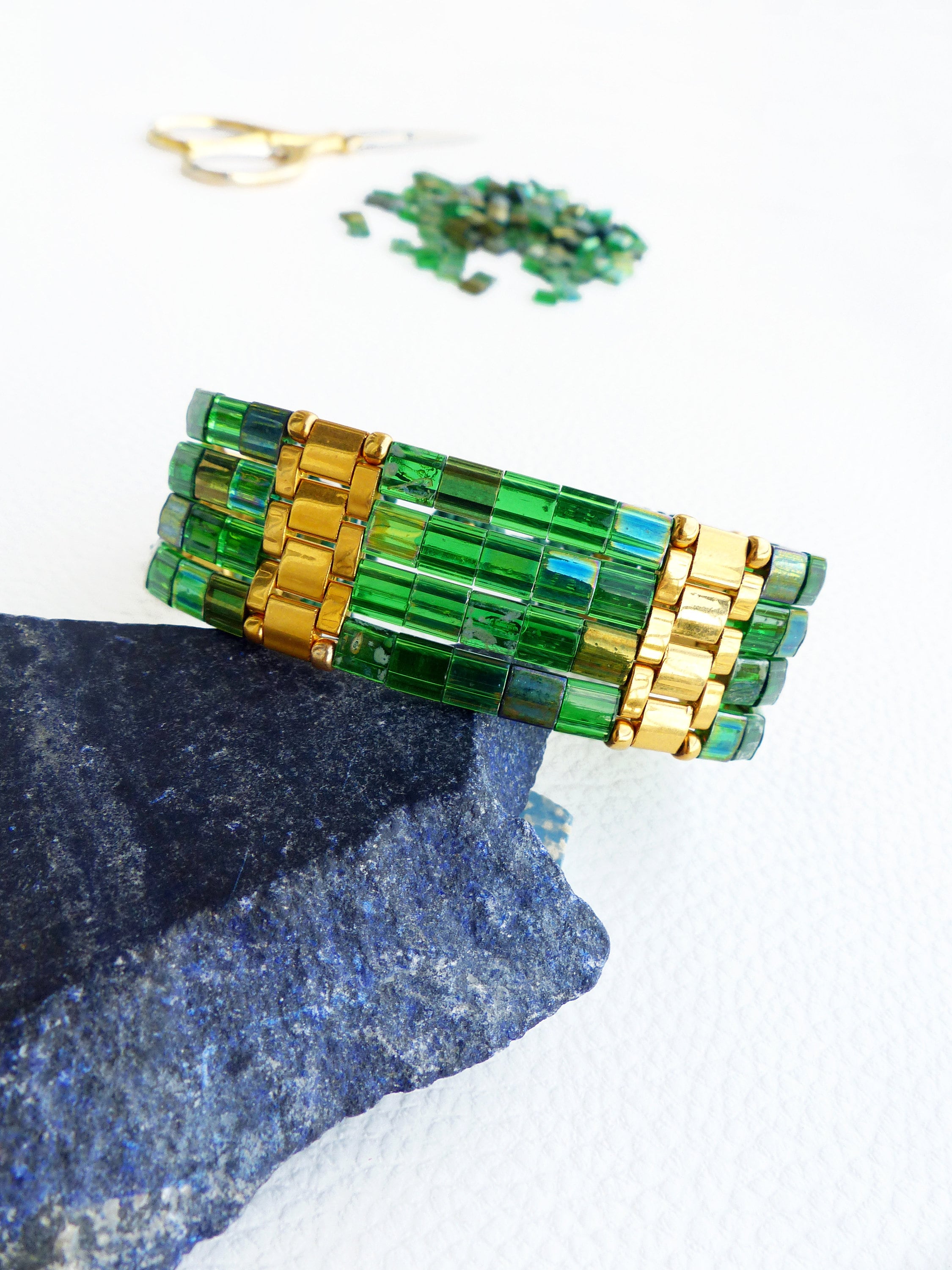 Peut inclure: Un bracelet en perles vertes et dorées avec un motif géométrique. Le bracelet est composé de petites perles carrées et est empilé sur lui-même.