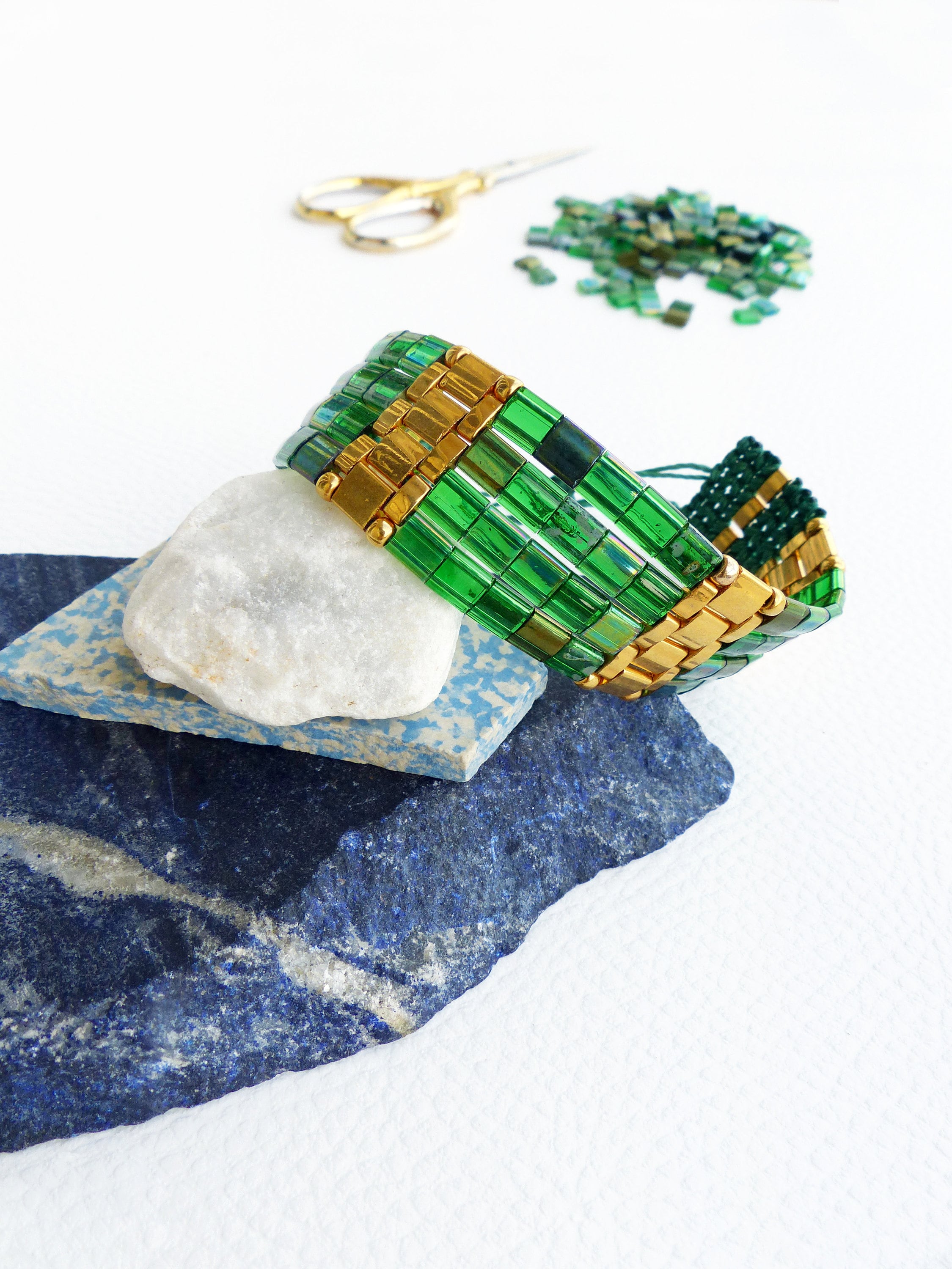 Peut inclure: Un bracelet en perles vertes et dorées avec un motif tissé. Le bracelet est posé sur une pile de pierres bleues et blanches.