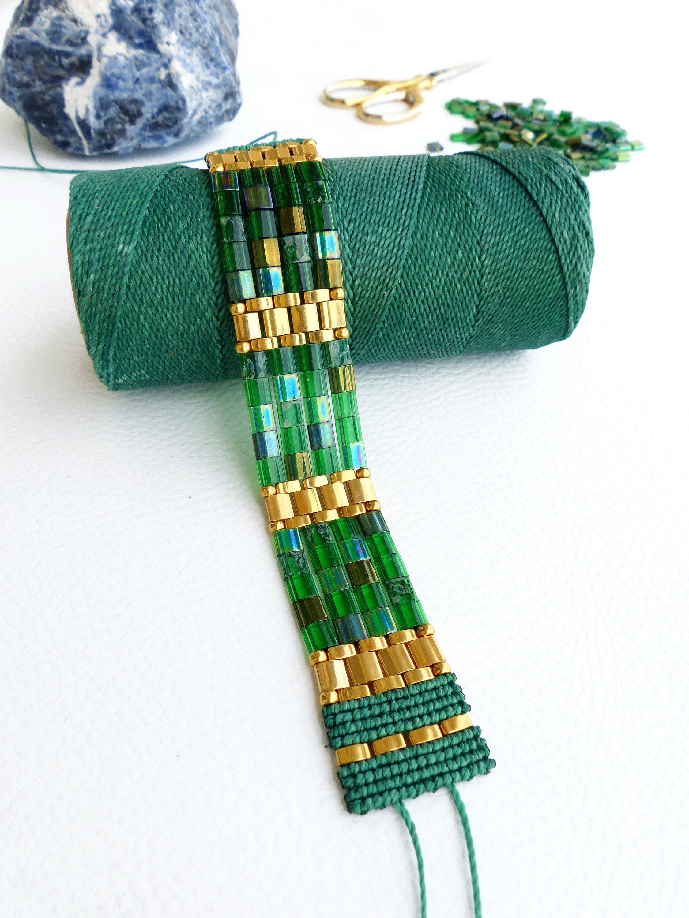 Peut inclure: Un bracelet en perles vertes et dorées est en cours de fabrication avec du fil vert. Le bracelet est composé de petites perles carrées et de perles dorées. Le fil est enroulé autour d'une bobine de fil vert.