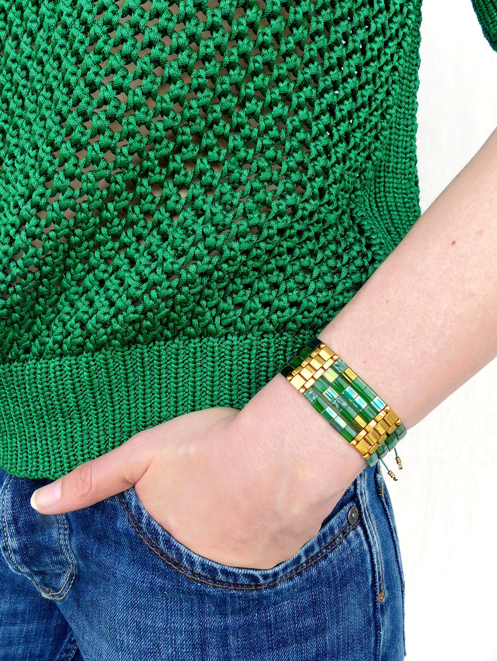 Peut inclure: Un bracelet en perles vert et or avec un design superposé. Le bracelet est composé de petites perles carrées dans des tons de vert et d'or, et est noué avec un cordon fin en or.