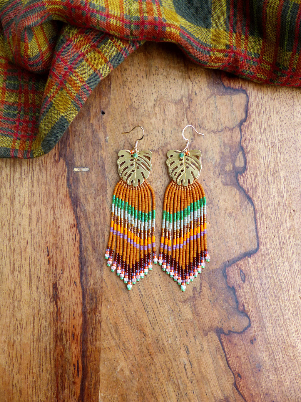 Peut inclure: Une paire de boucles d'oreilles pendantes en perles avec un motif de feuille dorée. Les boucles d'oreilles présentent un mélange de perles orange, vertes, violettes et marron. Les boucles d'oreilles sont présentées sur une surface en bois avec une écharpe à carreaux en arrière-plan.