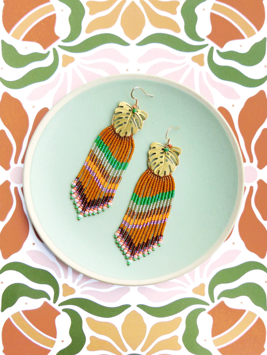 Peut inclure: Une paire de boucles d'oreilles à franges perlées avec un motif de feuille dorée. Les boucles d'oreilles présentent un mélange de perles orange, vertes, violettes et blanches. Elles sont présentées sur une assiette bleu clair.