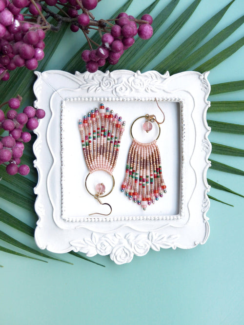 Peut inclure: Une paire de boucles d'oreilles en perles roses et dorées avec un design en frange. Les boucles d'oreilles sont présentées dans un cadre blanc à motif floral.