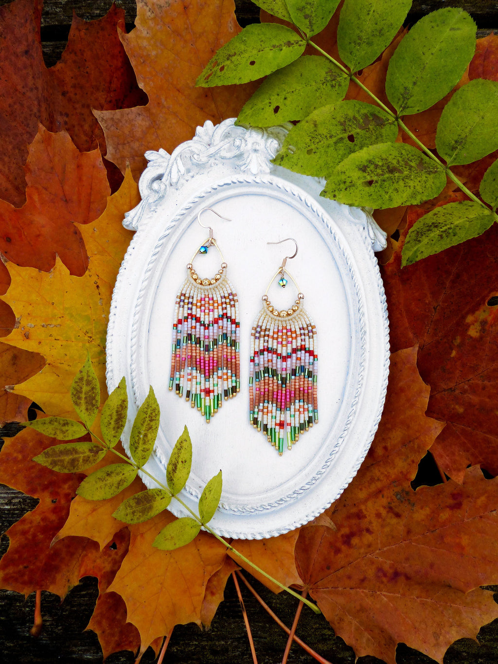 Peut inclure: Une paire de boucles d'oreilles en perles colorées avec un schéma de couleurs blanc, rose, vert et or. Les boucles d'oreilles sont suspendues à un cadre ovale blanc sur un fond de feuilles d'automne.