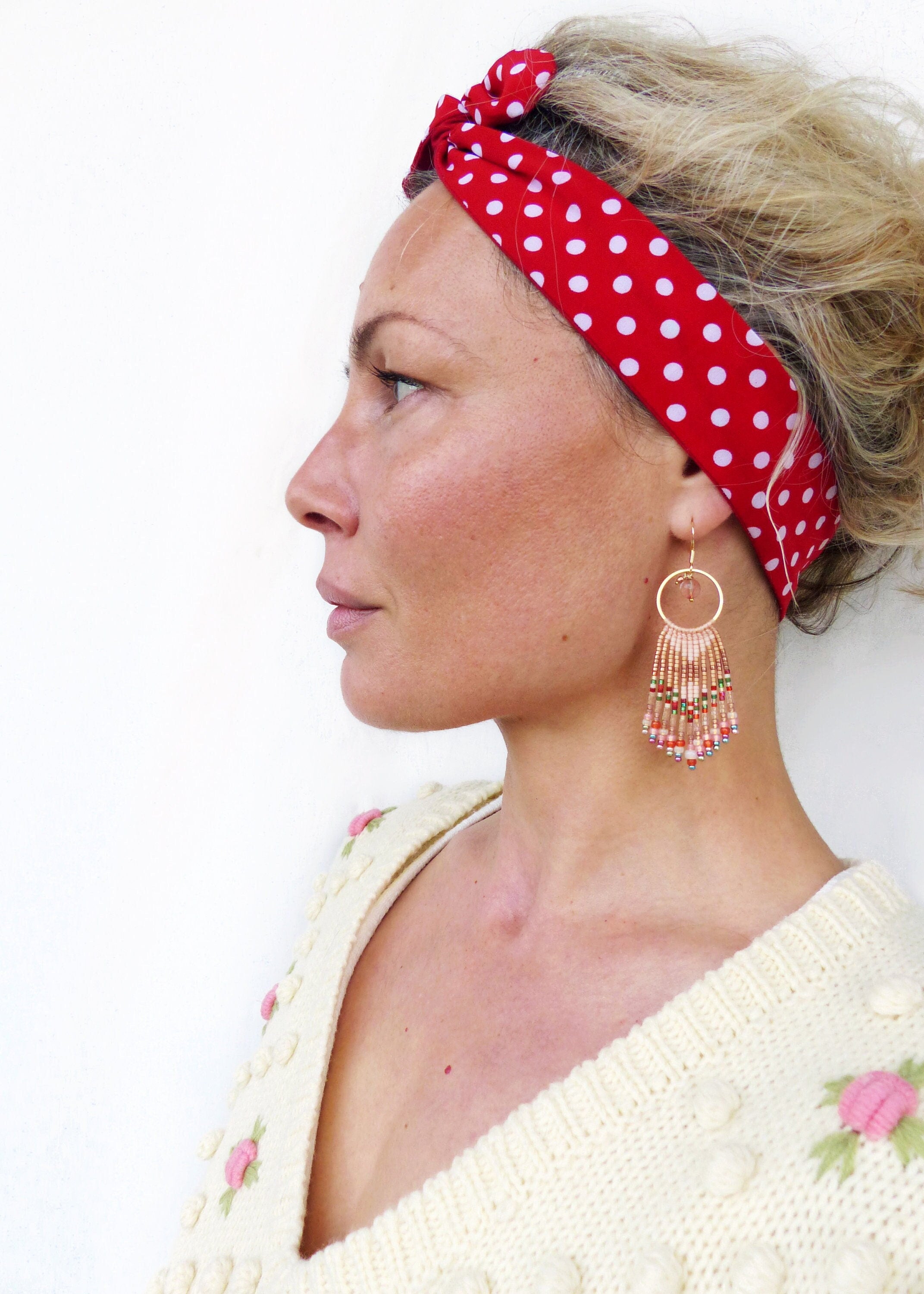 Peut inclure: Une femme portant un bandeau rouge à pois blancs et un pull crème avec des fleurs brodées roses. Elle porte de longues boucles d'oreilles en perles avec un anneau en or et une variété de perles colorées.