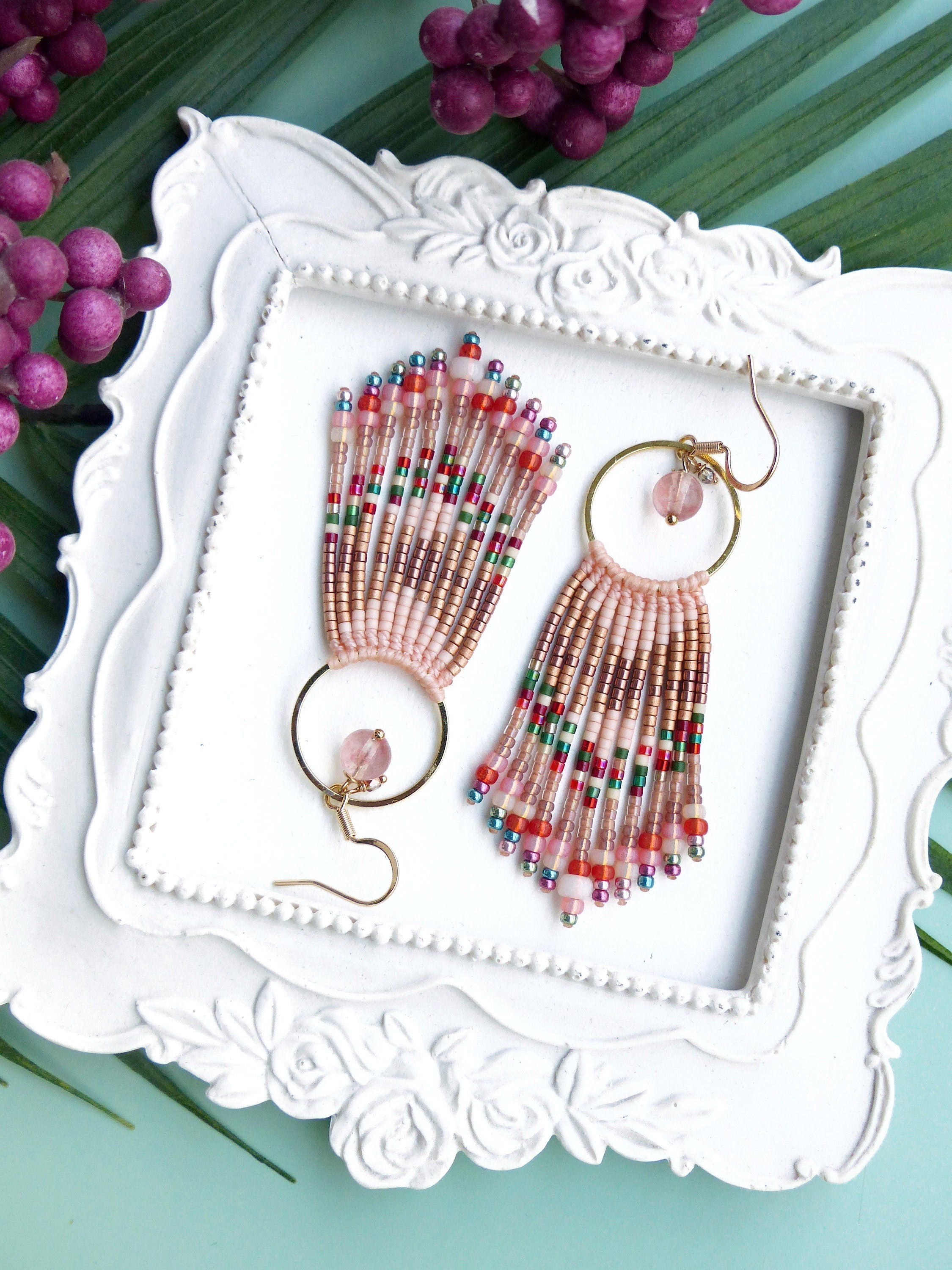 Peut inclure: Une paire de boucles d'oreilles en perles roses avec des cercles dorés et des accents de pierres roses. Les boucles d'oreilles sont présentées sur un plateau blanc à motif floral.