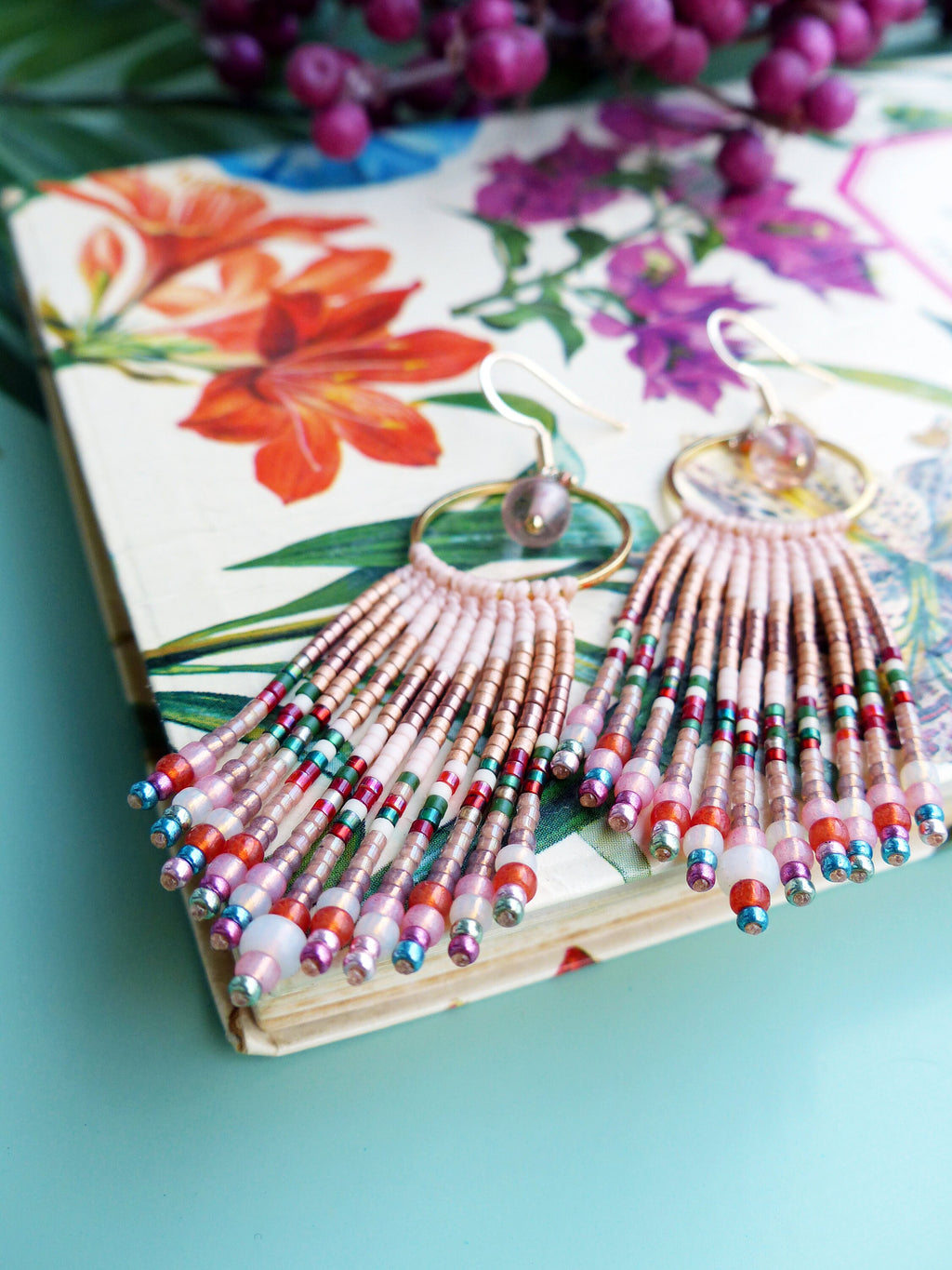 Peut inclure: Une paire de boucles d'oreilles en forme de pompon en perles roses et dorées avec une variété de couleurs, notamment le rouge, le vert et le bleu. Les boucles d'oreilles sont suspendues à une surface à motifs floraux.