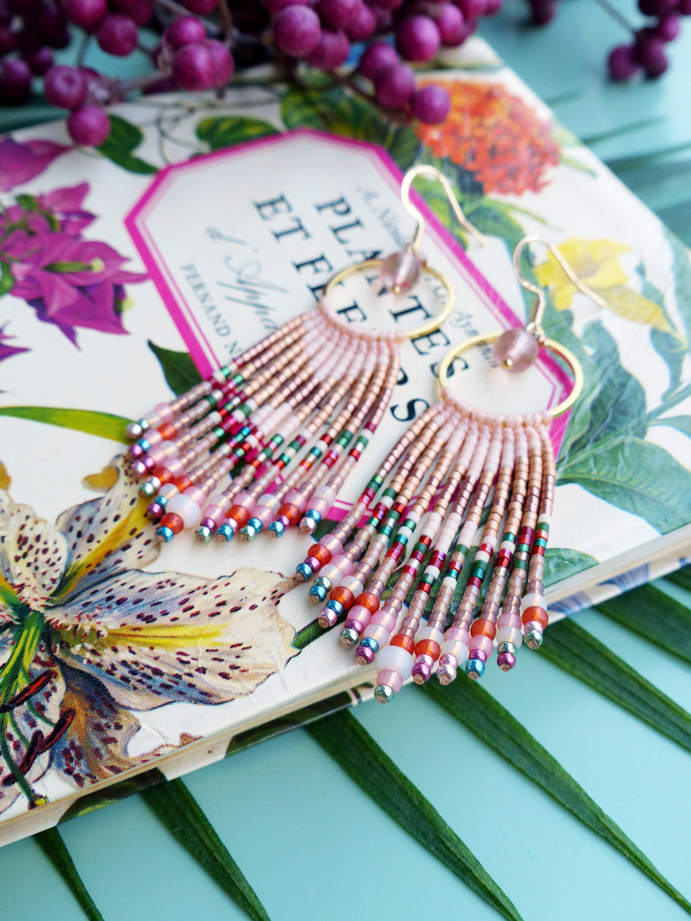 Peut inclure: Une paire de boucles d'oreilles en franges de perles roses et dorées avec un design de cerceau. Les boucles d'oreilles sont sur un livre avec un motif floral et le texte "Plantes et Fleurs".