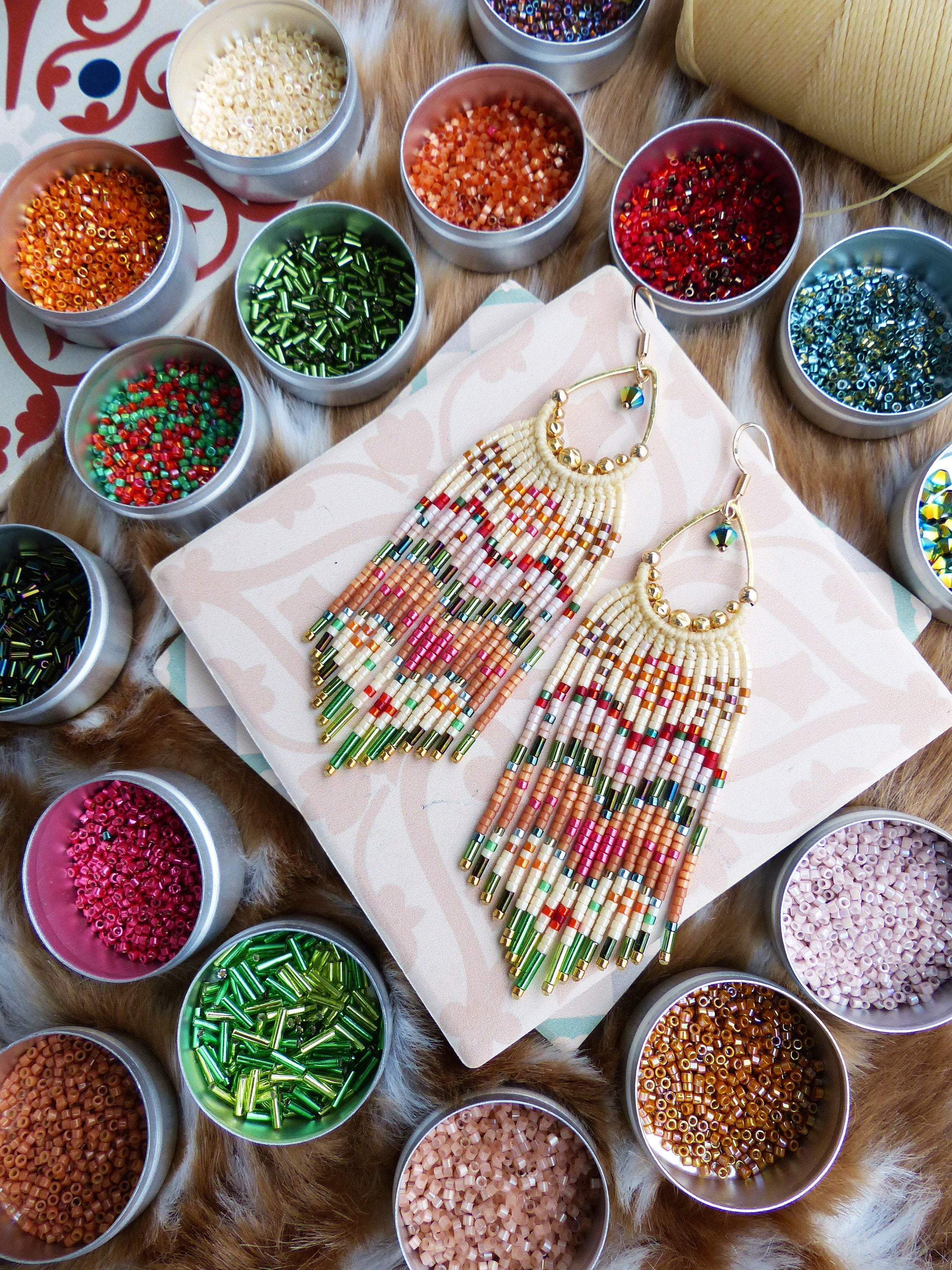 Peut inclure: Une paire de longues boucles d'oreilles en perles avec un motif géométrique dans des tons de rose, vert, blanc et marron. Les boucles d'oreilles sont suspendues à des créoles dorées et sont entourées de petits contenants de perles colorées.