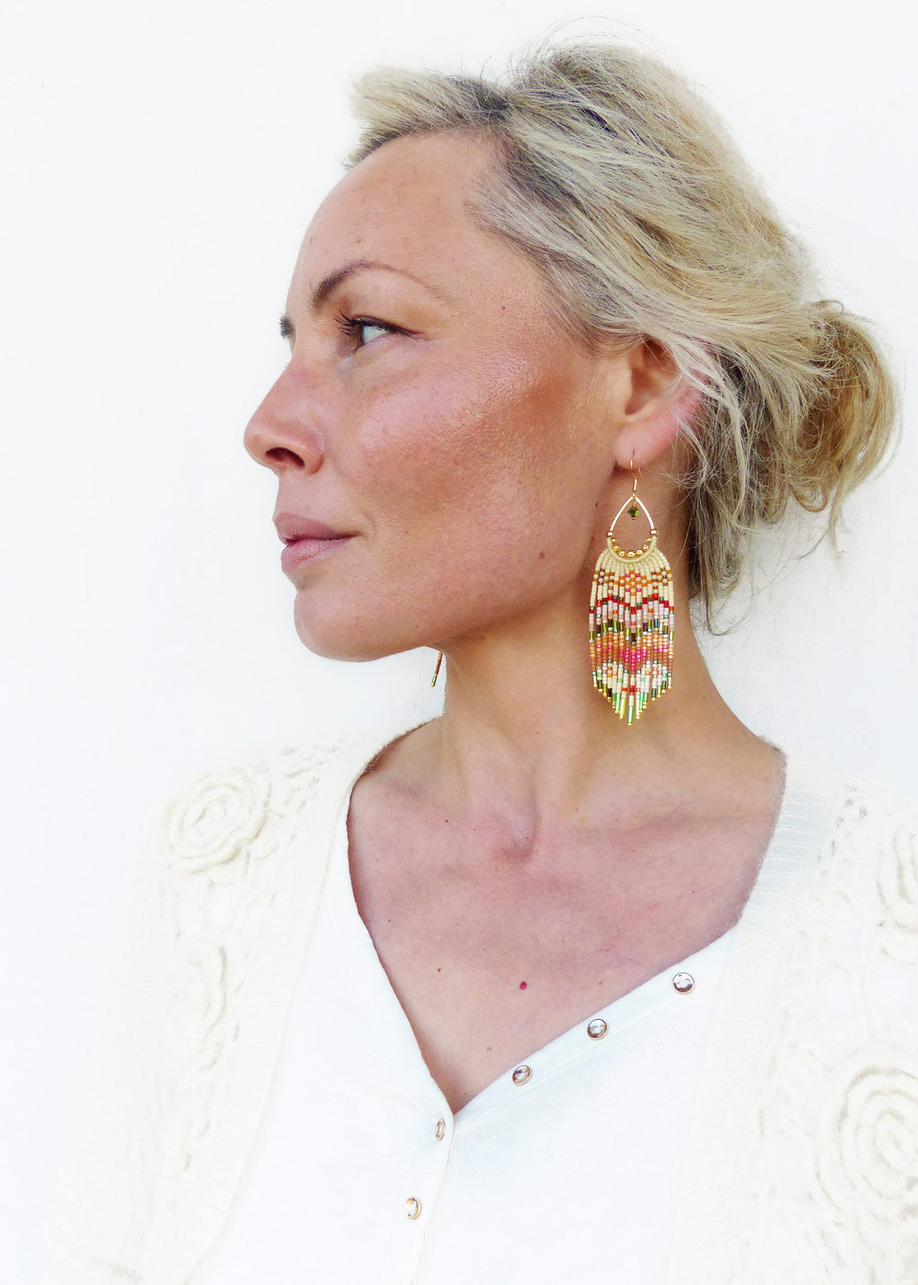 Peut inclure: Une femme portant un cardigan en crochet blanc et une chemise blanche à boutons. Elle porte de longues boucles d'oreilles en perles avec un motif géométrique dans des tons de blanc, orange, rose, vert et or.