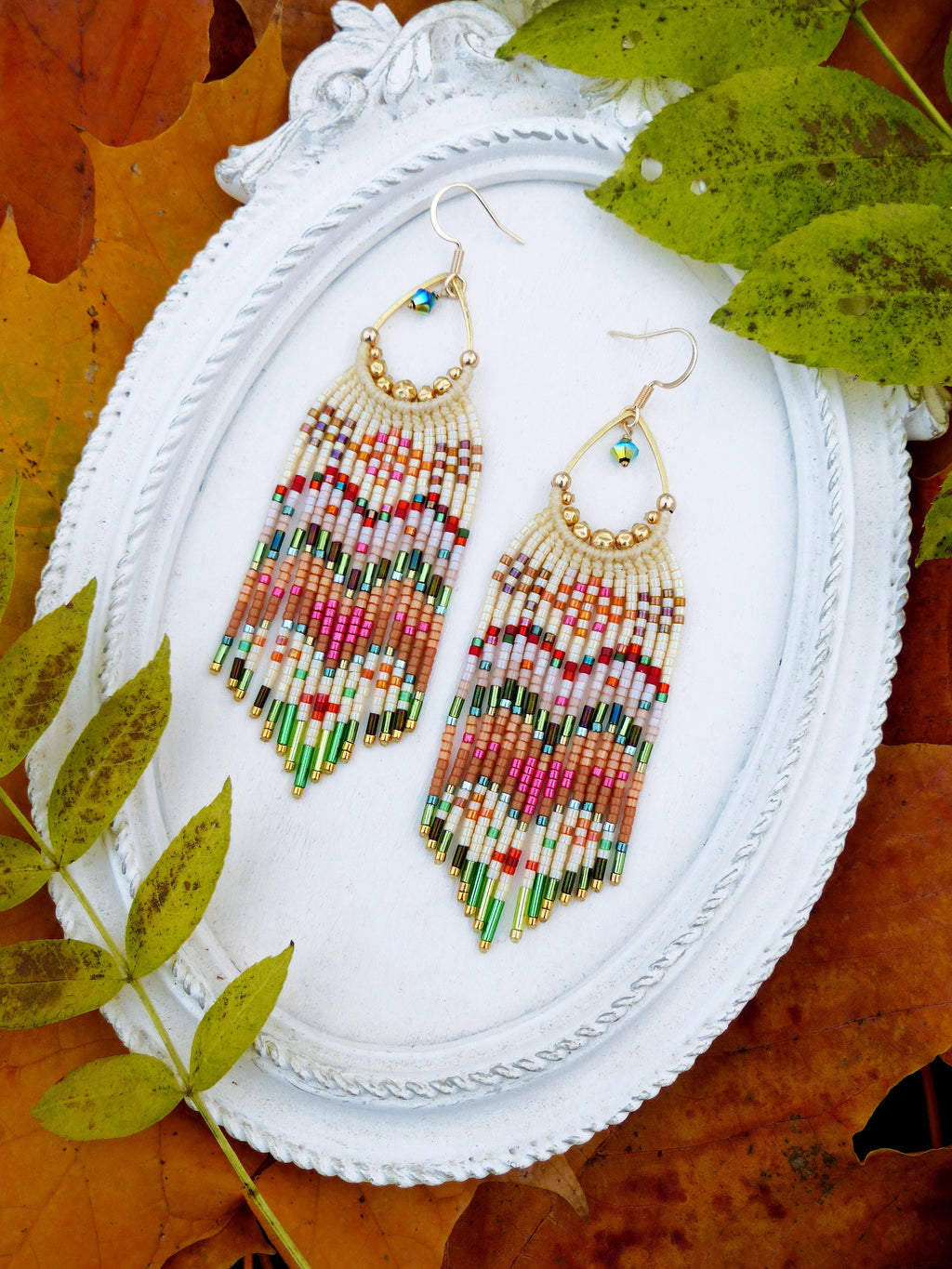 Peut inclure: Une paire de longues boucles d'oreilles en perles colorées avec des accents dorés. Les boucles d'oreilles sont suspendues à un cadre blanc de forme ovale.