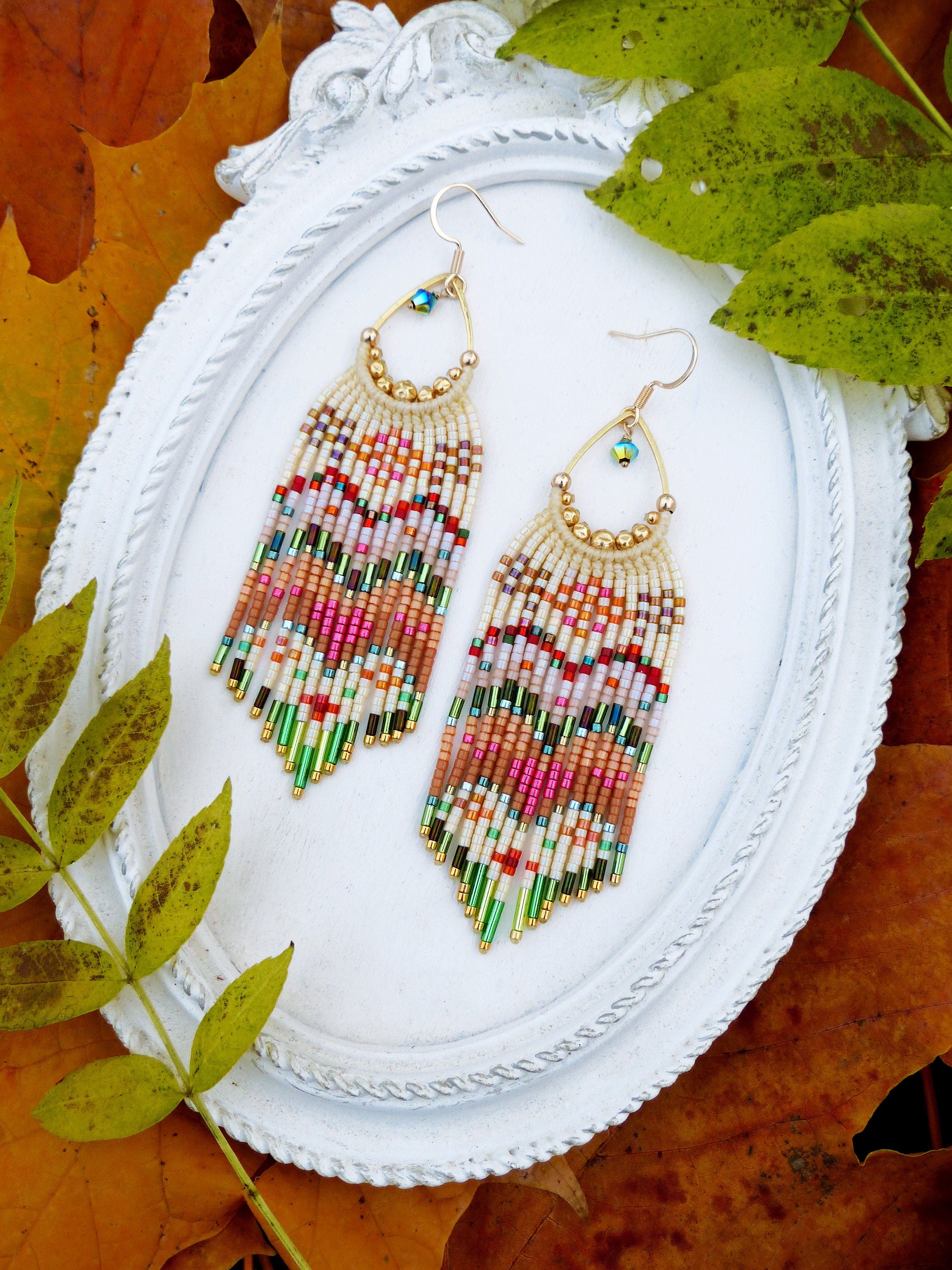 Peut inclure: Une paire de longues boucles d'oreilles en perles colorées avec des accents dorés. Les boucles d'oreilles sont suspendues à un cadre blanc de forme ovale.