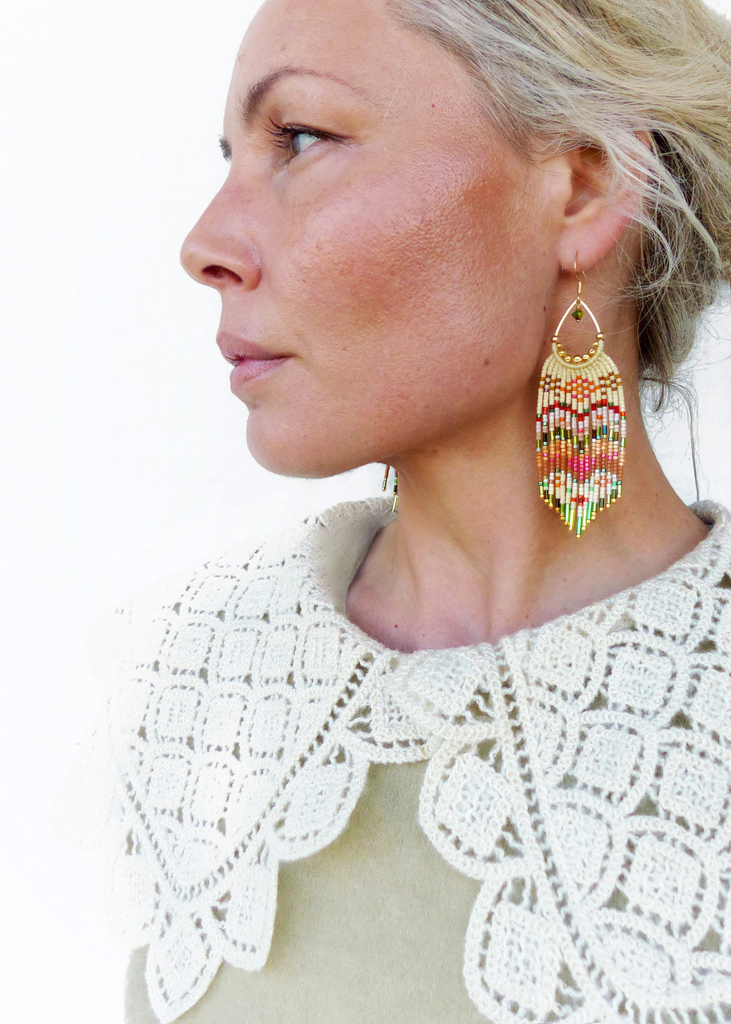Peut inclure: Une paire de longues boucles d'oreilles en perles colorées avec un crochet doré. Les boucles d'oreilles sont faites avec une variété de perles, y compris le blanc, le rose, l'orange, le vert et l'or. Les boucles d'oreilles pendent des lobes d'oreilles d'une personne portant un col en crochet blanc.